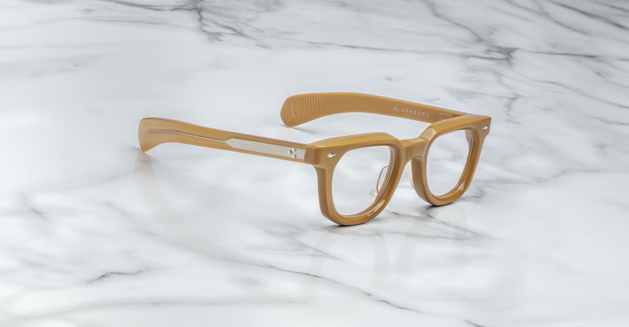 A pair of Jacques Marie Mage Vendome optical frames by Jacques Marie Mage, featuring thick tan rims and clear lenses, handcrafted in Japan, rests on a white marble surface.