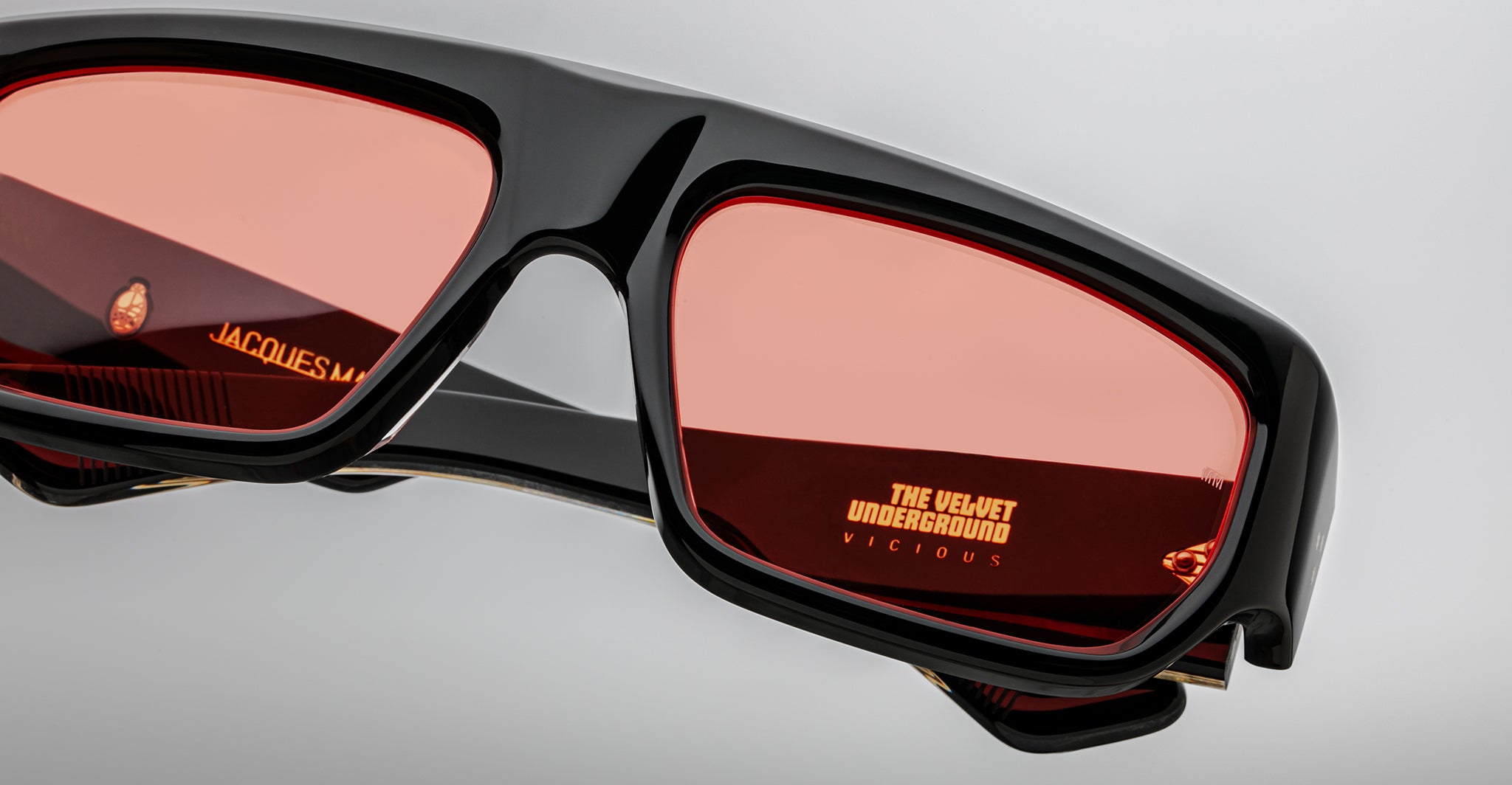 Close-up of Jacques Marie Mage Vicious black sunglasses with red lenses. The left lens reflects “JACQUES,” while the right lens features “THE VELVET UNDERGROUND VICIOUS” in yellow text.