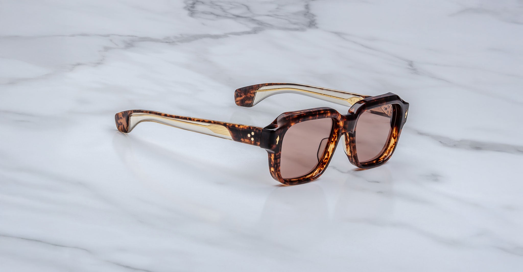 A pair of square, tortoiseshell sunglasses with brown-tinted lenses rests on a white marble surface with grey veins.