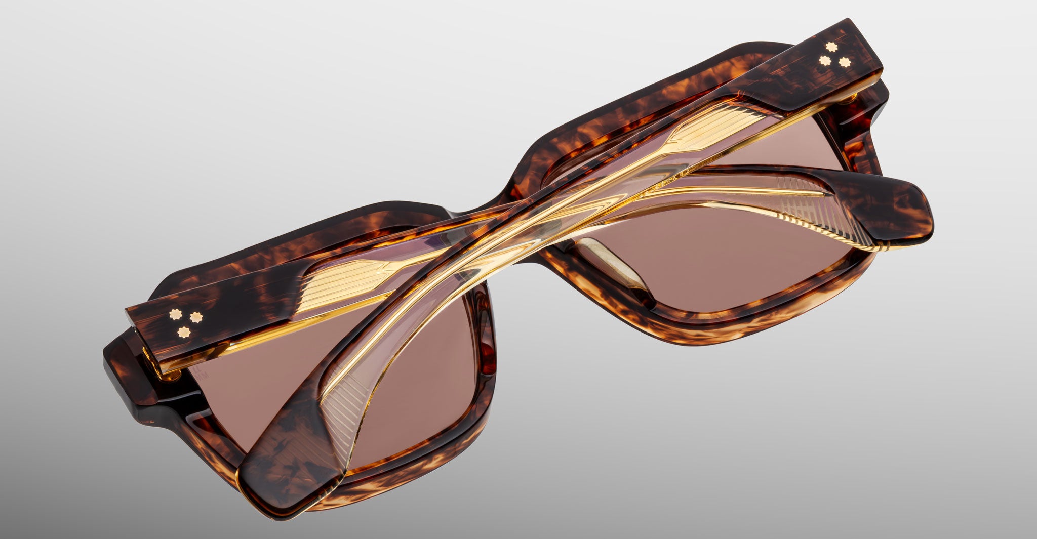 A pair of tortoiseshell sunglasses with tinted lenses, folded and viewed from above. The arms feature gold accents and crisscross over each other, set against a light gray background.