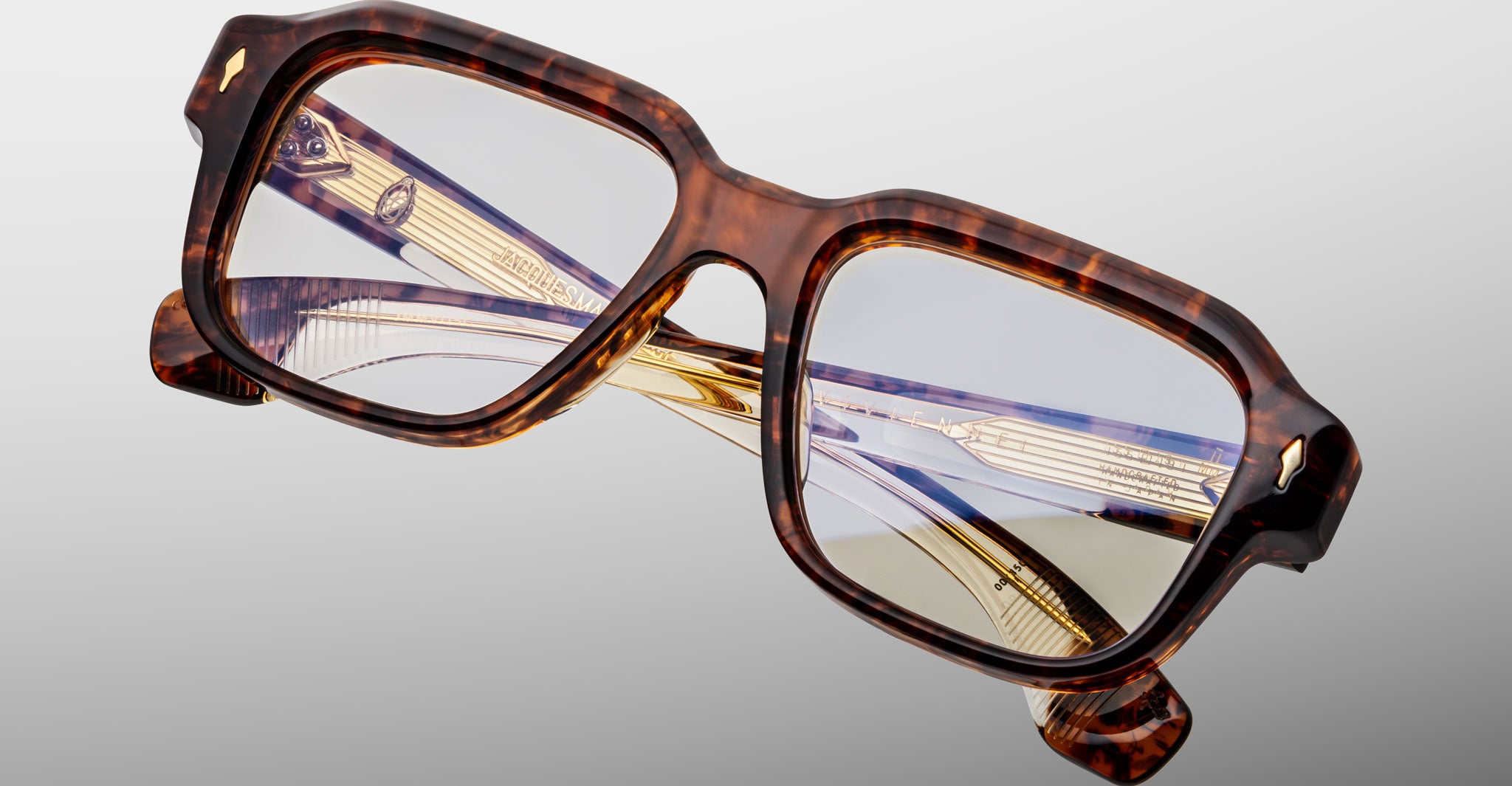 The Jacques Marie Mage Vivienne eyeglasses feature a bold, thick rectangular tortoiseshell frame with gold and clear decorative temples, displayed folded on a light gray background.