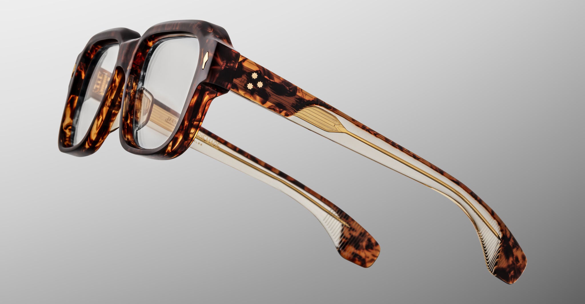The Jacques Marie Mage Vivienne eyeglasses feature thick, square tortoiseshell frames and clear lenses, shown from a diagonal angle against a plain gray background. Gold detailing and small decorative accents adorn the temples.