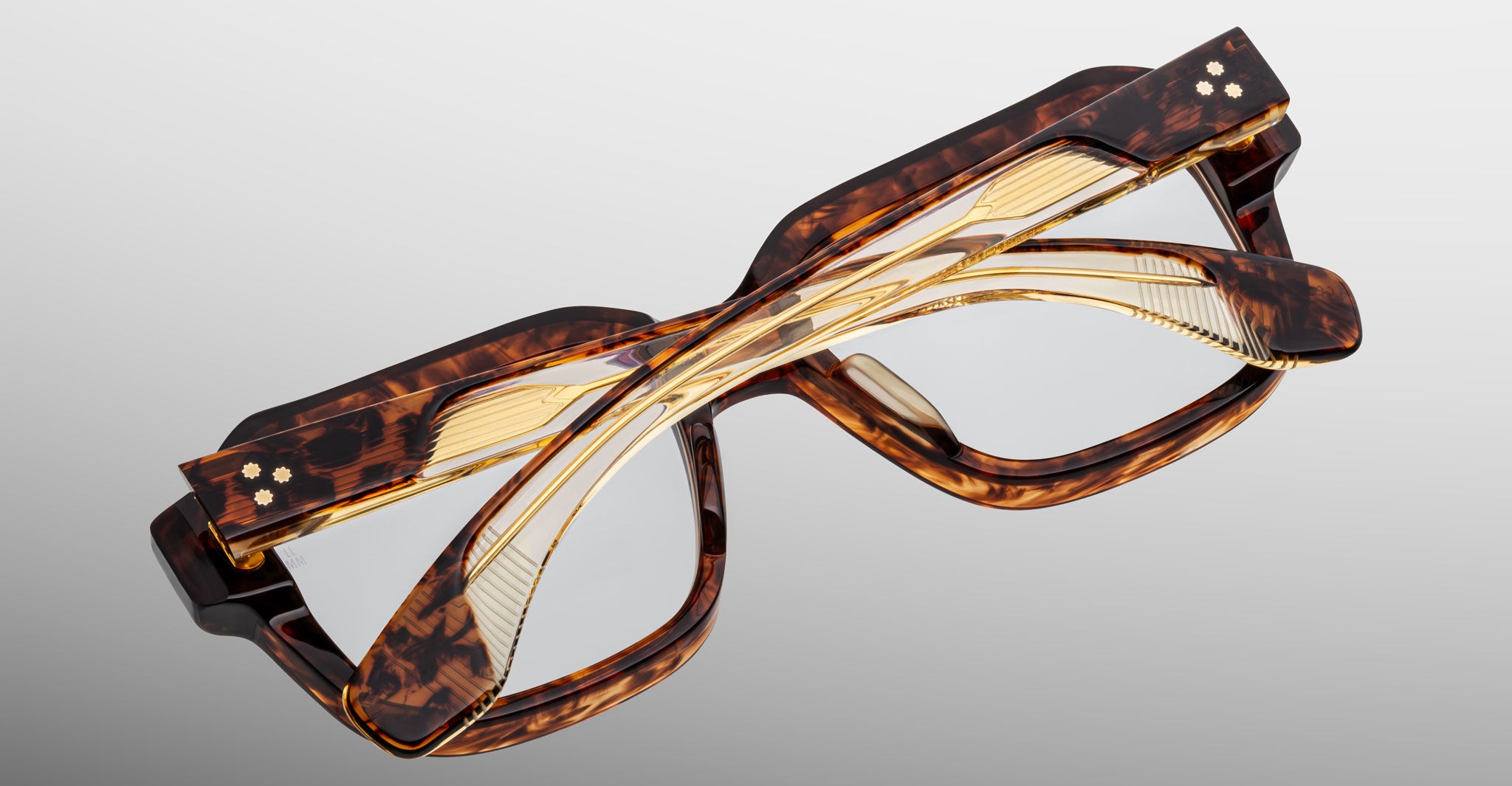 The Jacques Marie Mage Vivienne eyeglasses in tortoiseshell with gold accents are folded with crossed arms and displayed on a light gray background.