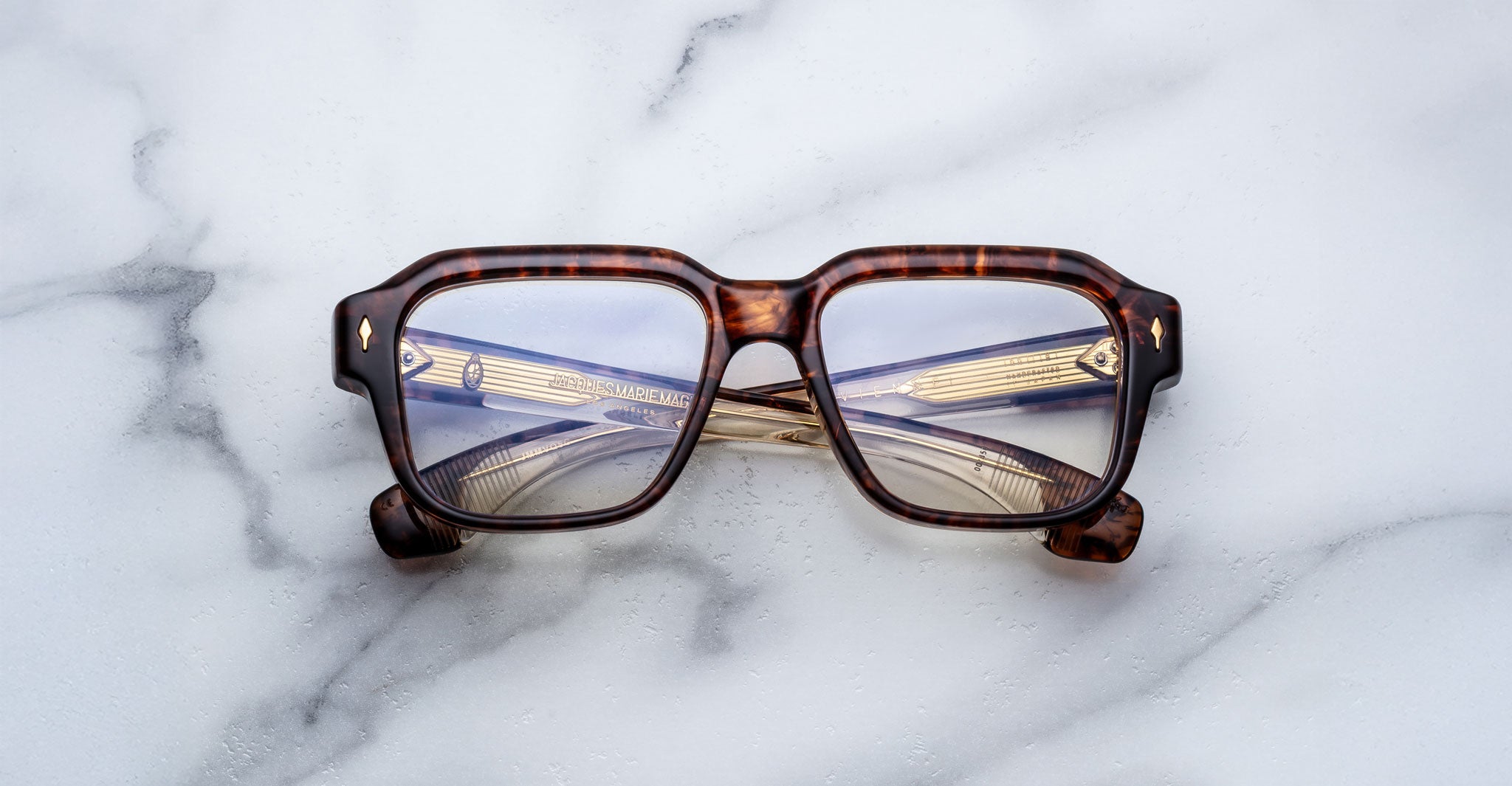 Jacques Marie Mage Vivienne square eyeglasses in brown tortoiseshell with gold decorative temples, folded and resting on a white marble surface.