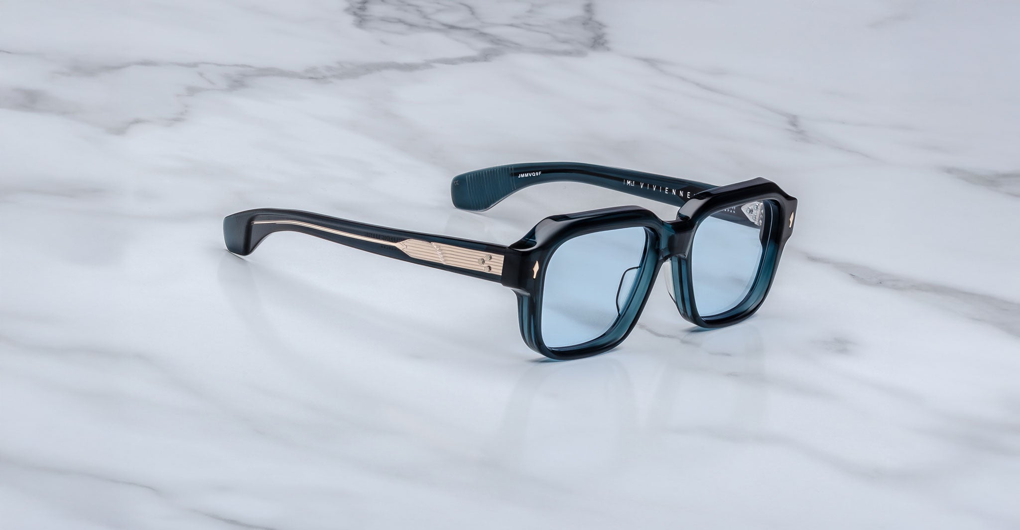 The Jacques Marie Mage Vivienne sunglasses feature blue lenses, square frames, and thick black arms with gold trim, elegantly displayed on a white marble surface.