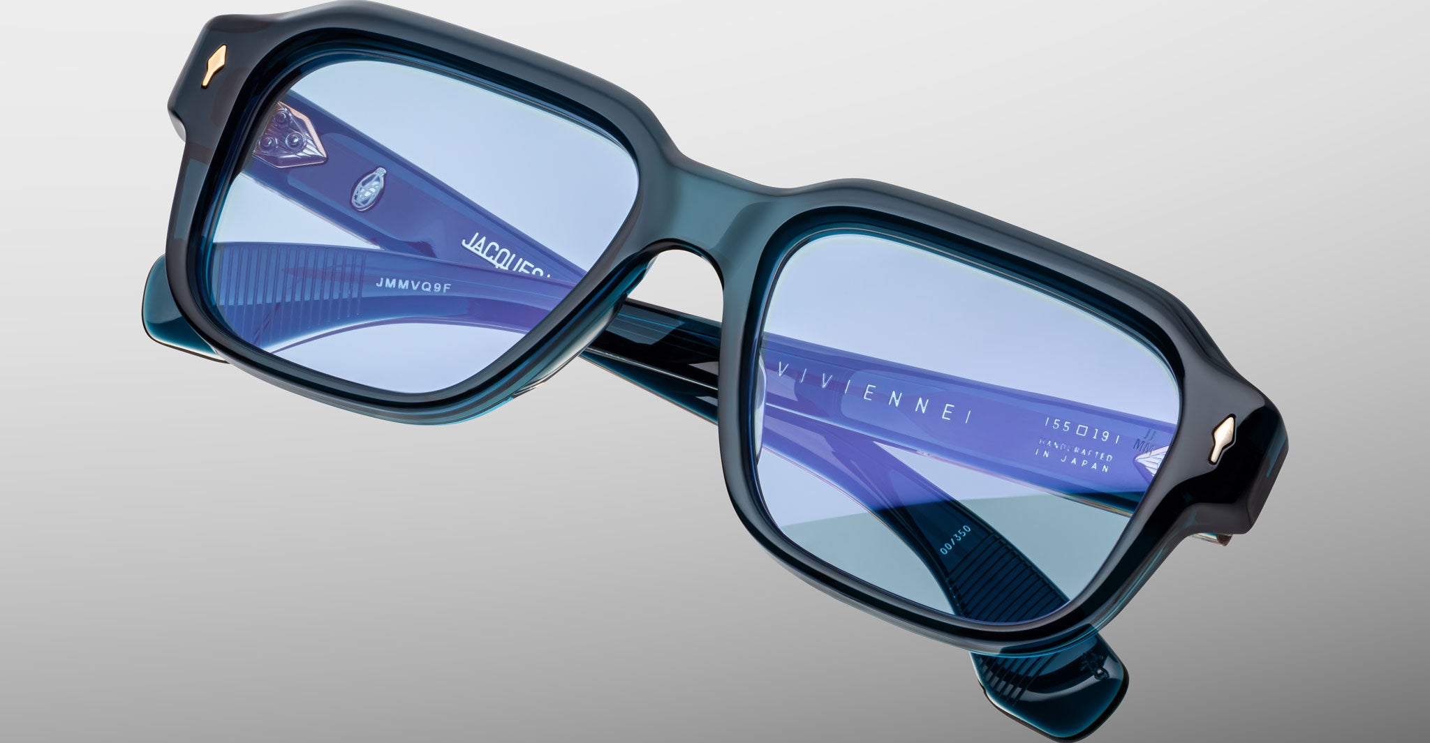 Close-up of Jacques Marie Mage Vivienne eyeglasses: stylish, rectangular blue-green frames with thick rims, blue-tinted lenses, and gold corner accents, displayed against a light gray background.