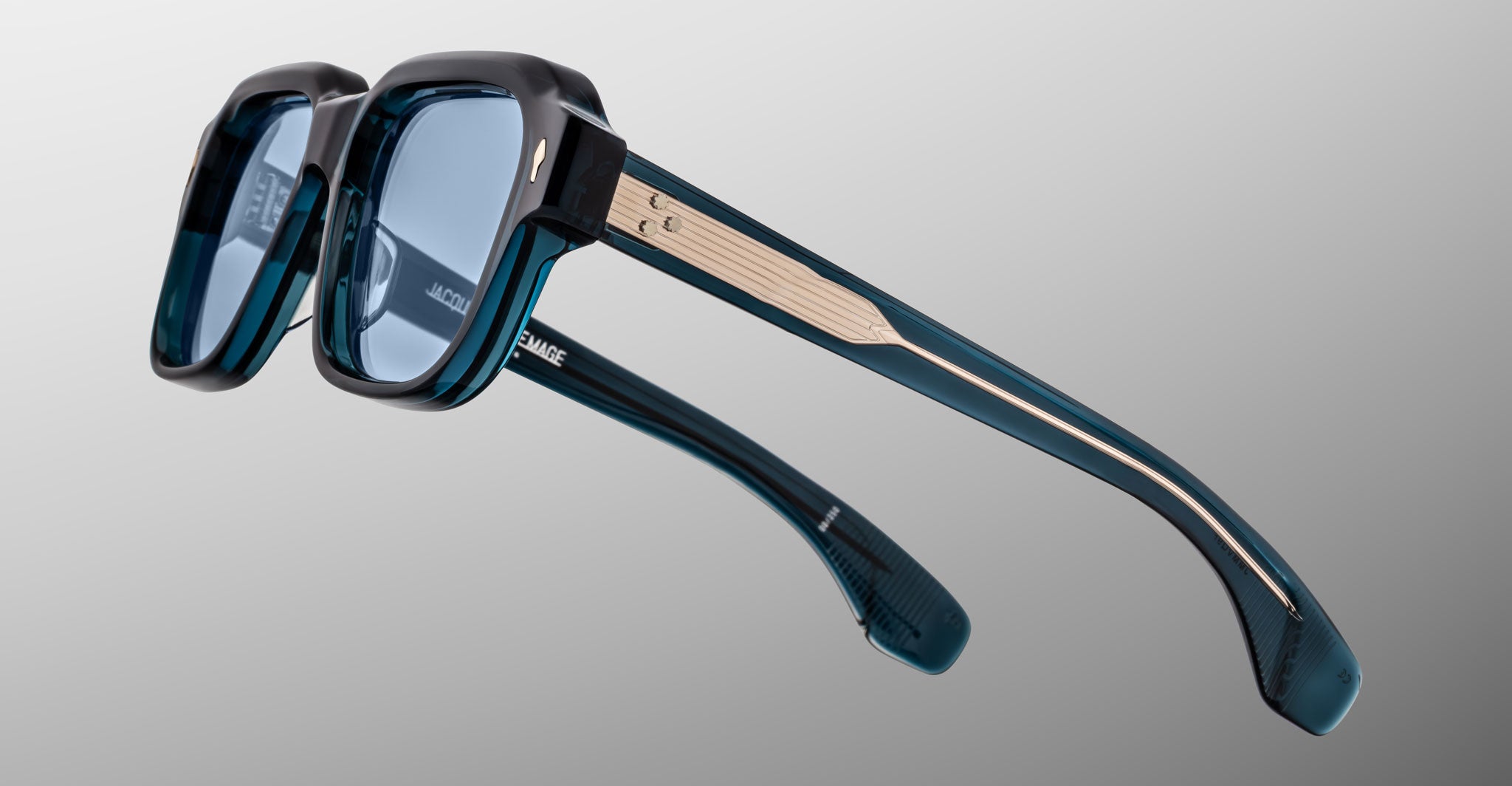Close-up of Jacques Marie Mage Vivienne sunglasses featuring blue-tinted square lenses, thick teal and black frames, metal arm accents, and a subtle logo inside the temple, set against a gradient gray background.