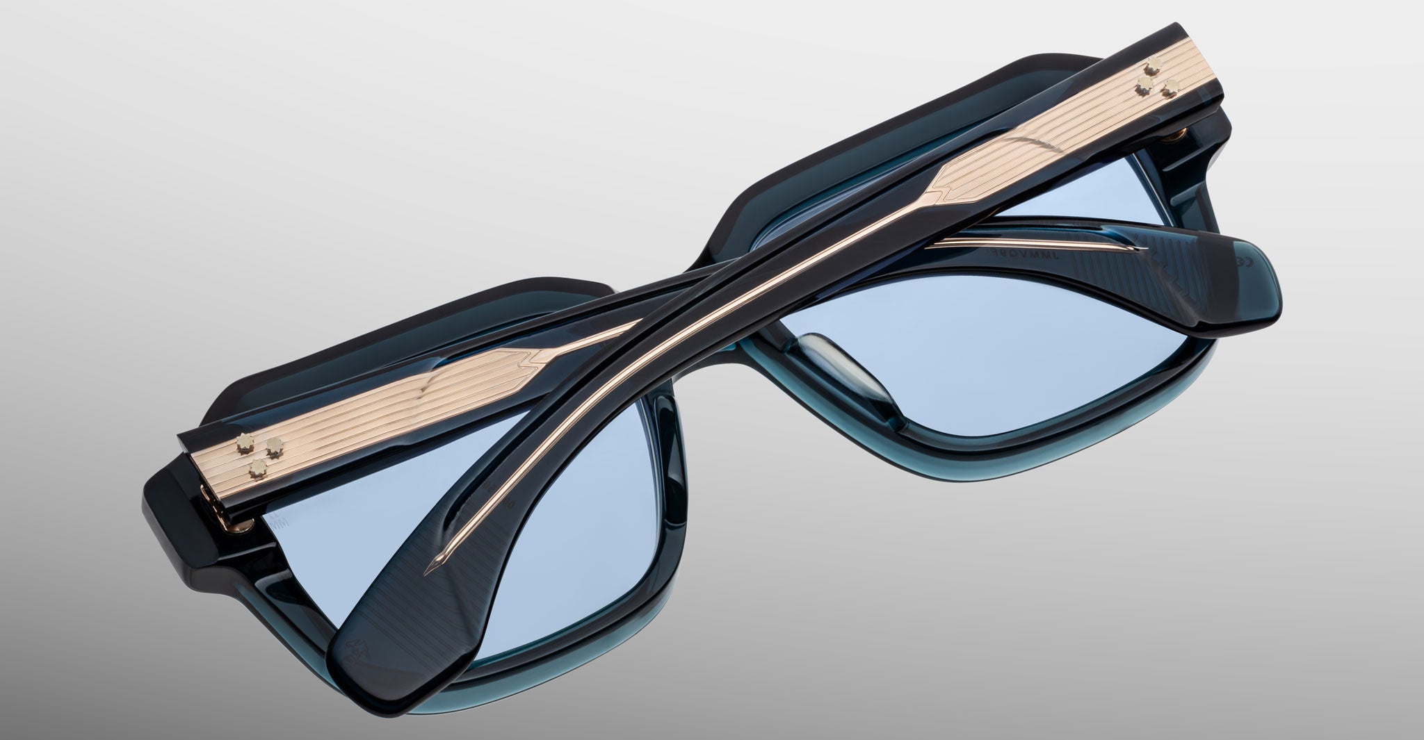 The Jacques Marie Mage Vivienne sunglasses feature blue-tinted lenses and gold-accented black arms, elegantly folded on a light gray gradient background.
