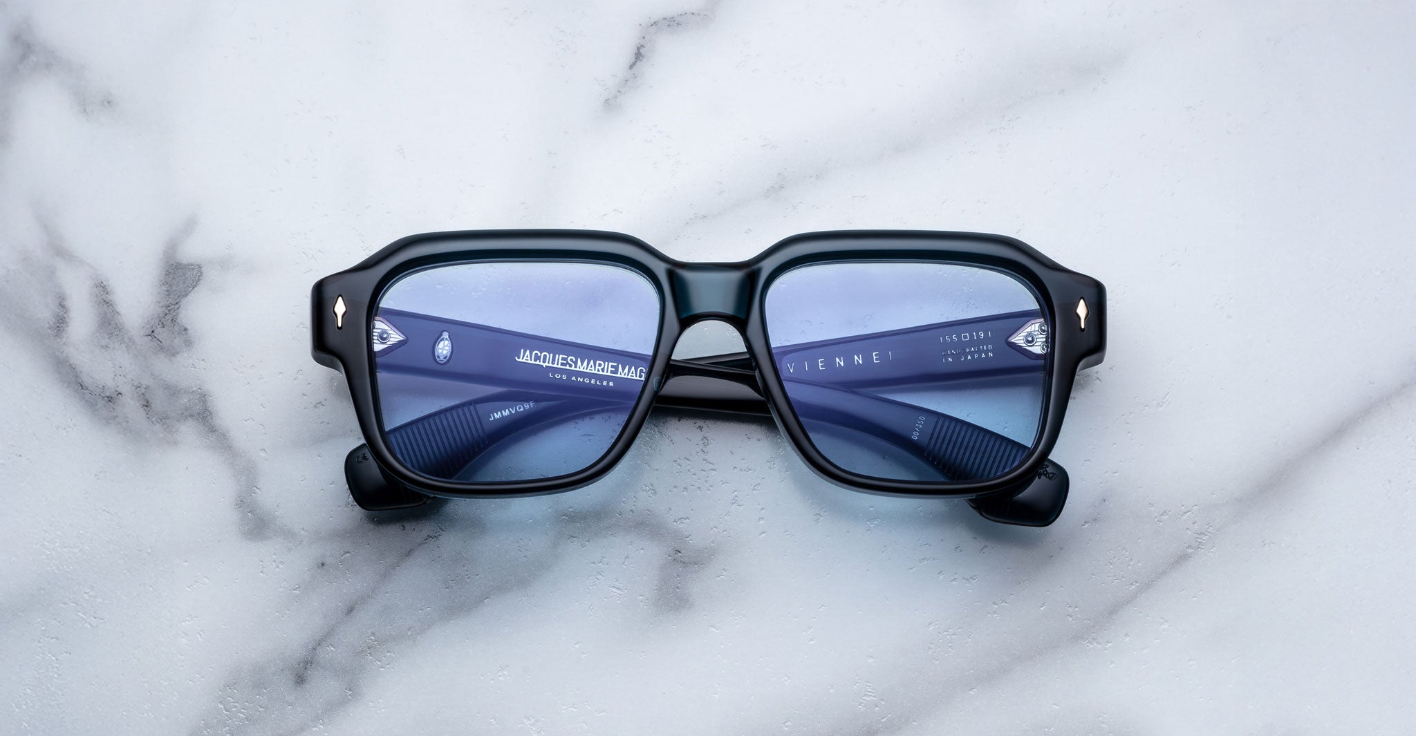 A pair of Jacques Marie Mage Vivienne square sunglasses with black frames and blue-tinted lenses rests on a white marble surface. Text on the inside of the left arm is visible through the lens.