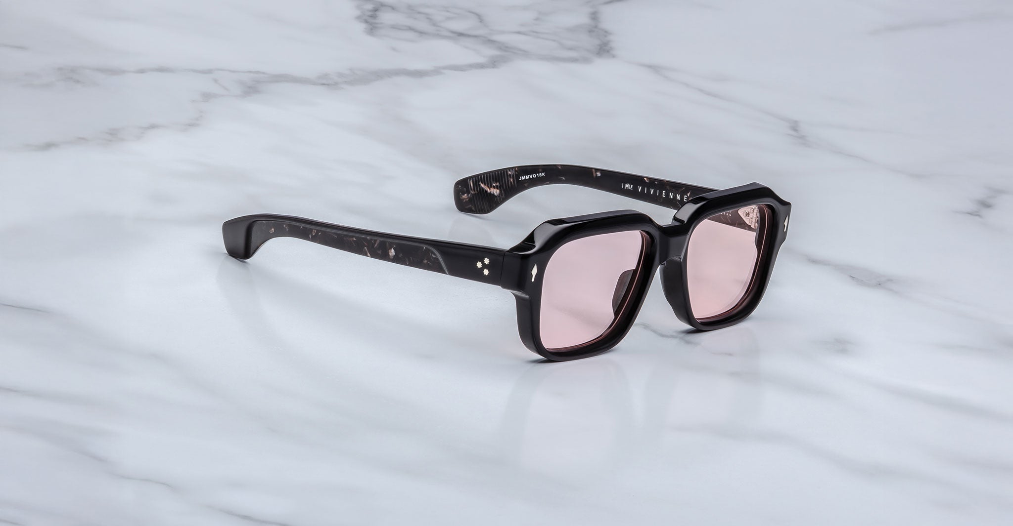 The Jacques Marie Mage Vivienne sunglasses feature a stylish black frame and pink-tinted lenses, elegantly displayed on a white marble surface.