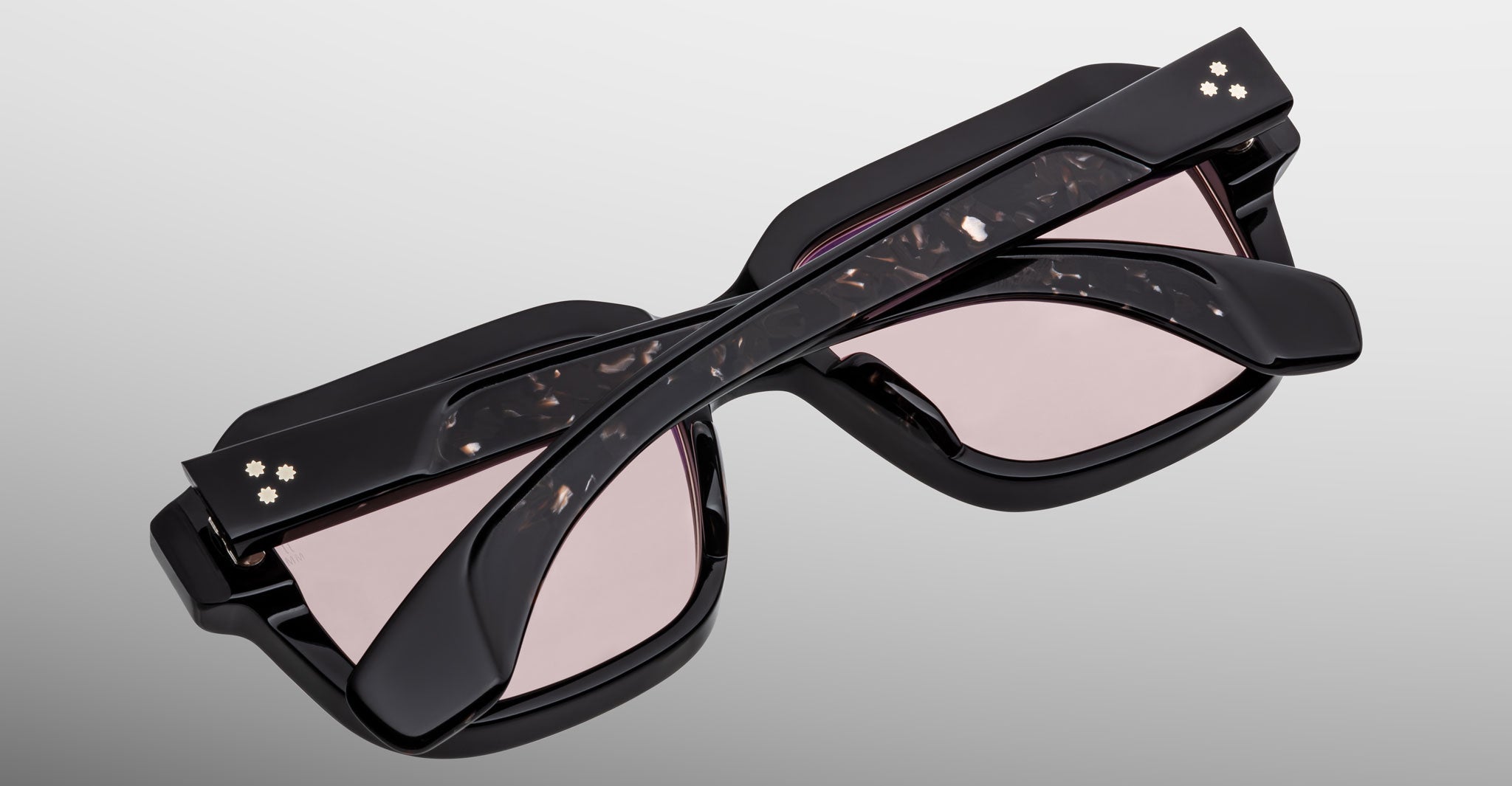 The Jacques Marie Mage Vivienne sunglasses feature a stylish black frame with light pink lenses, marbled temples, and gold accents near the hinges. Shown folded from above on a plain gradient light gray background.