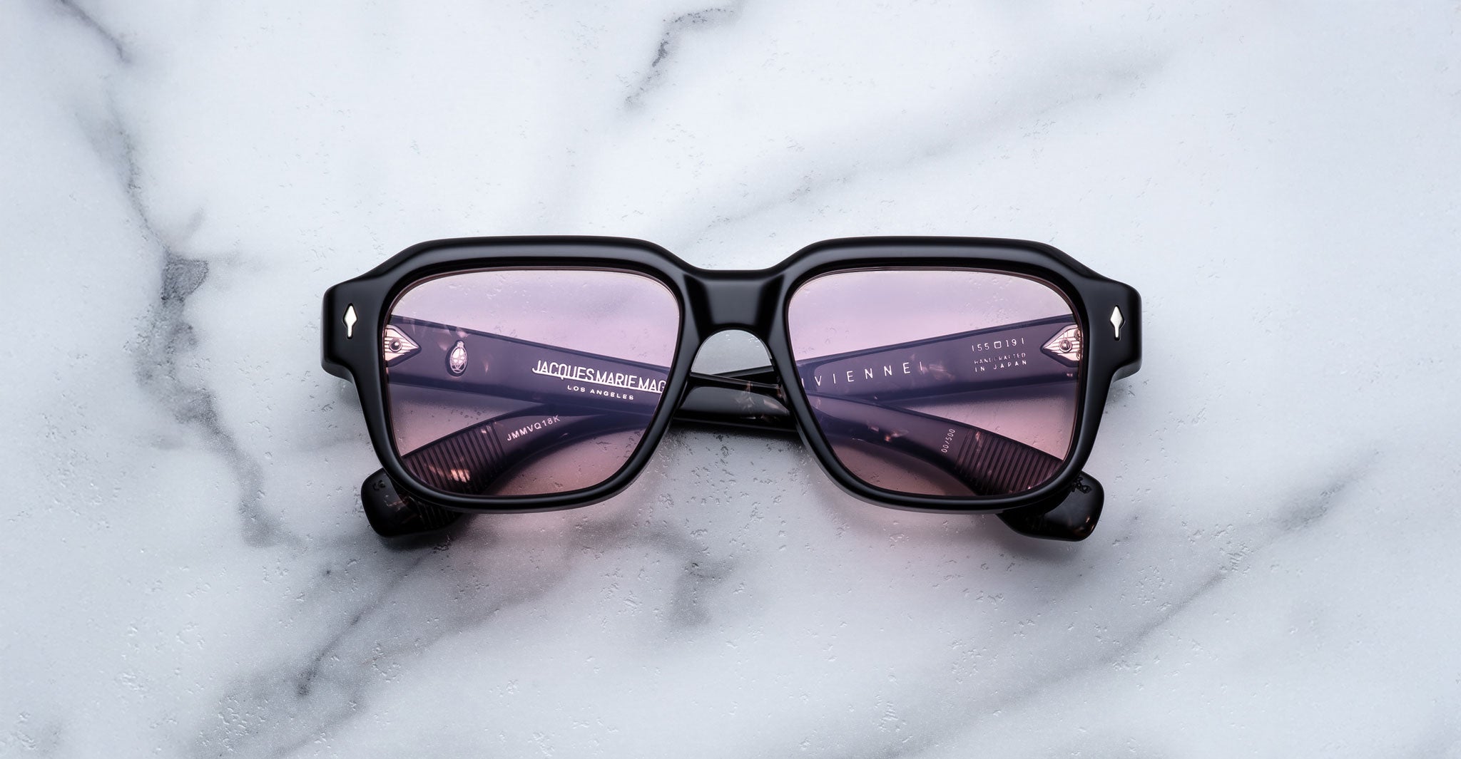 The Jacques Marie Mage Vivienne is a pair of black rectangular sunglasses with purple-tinted lenses, shown resting on a white marble surface.