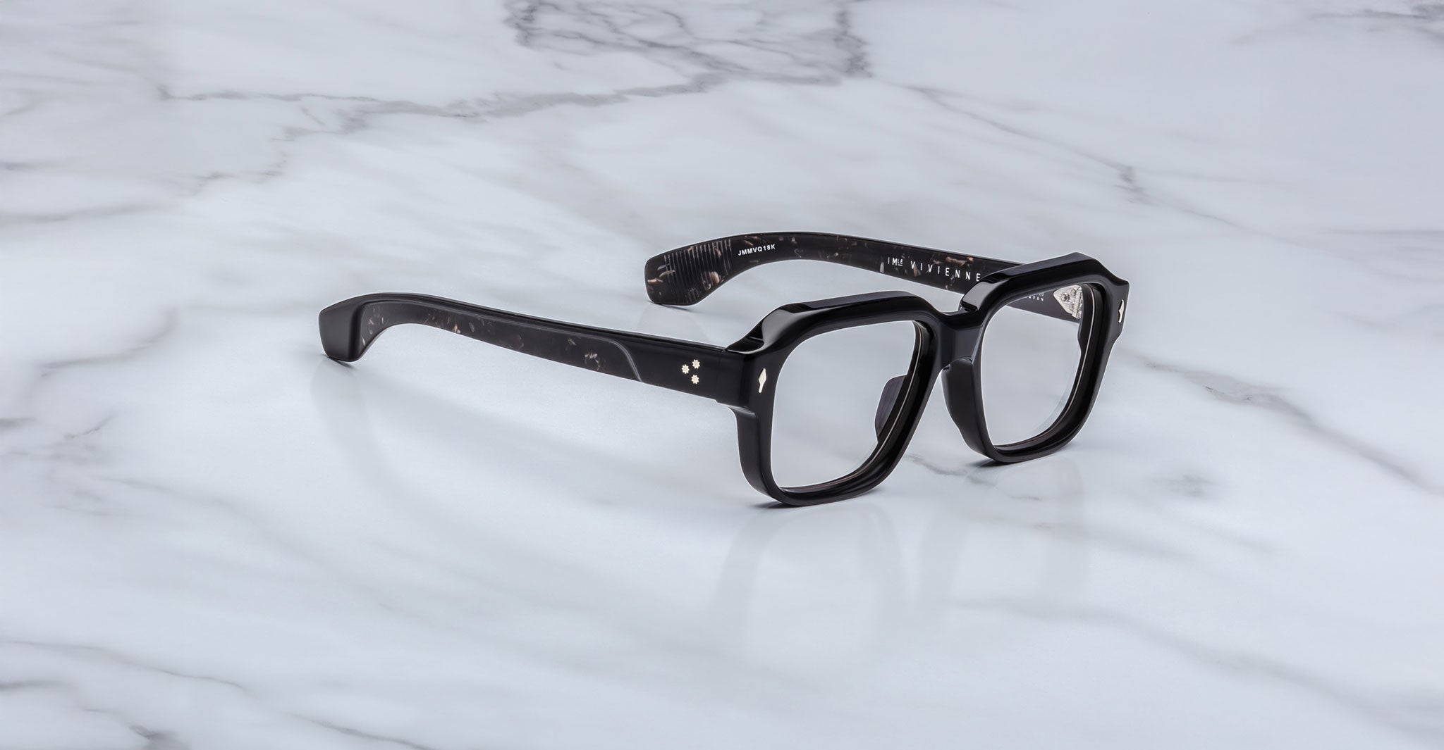 A pair of Jacques Marie Mage Vivienne black, thick-framed eyeglasses with clear lenses rests on a white marble surface.