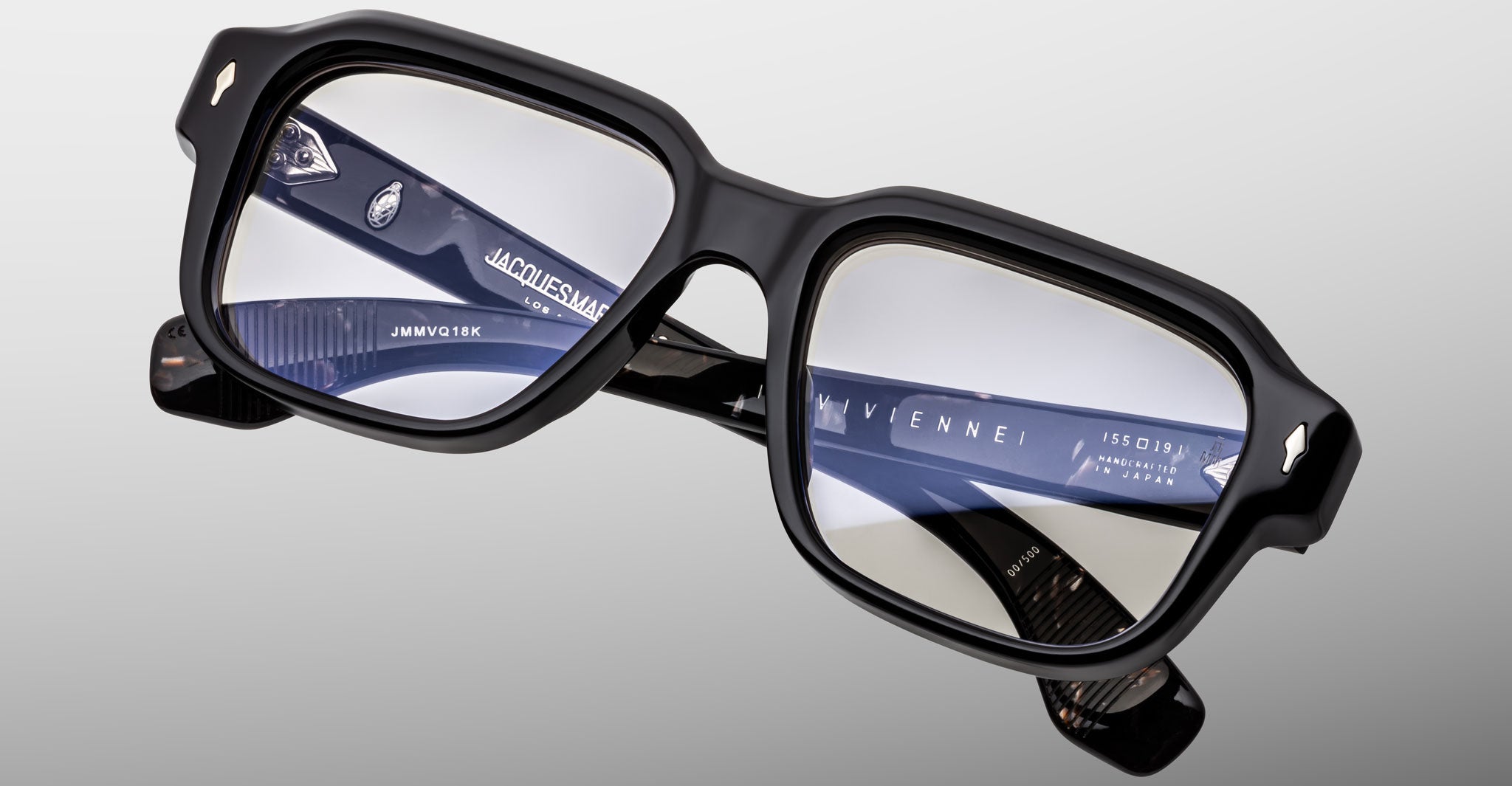 A close-up of Jacques Marie Mage Vivienne black square eyeglasses with thick frames and clear lenses, featuring model details engraved on the inner temples, set against a light gray background.