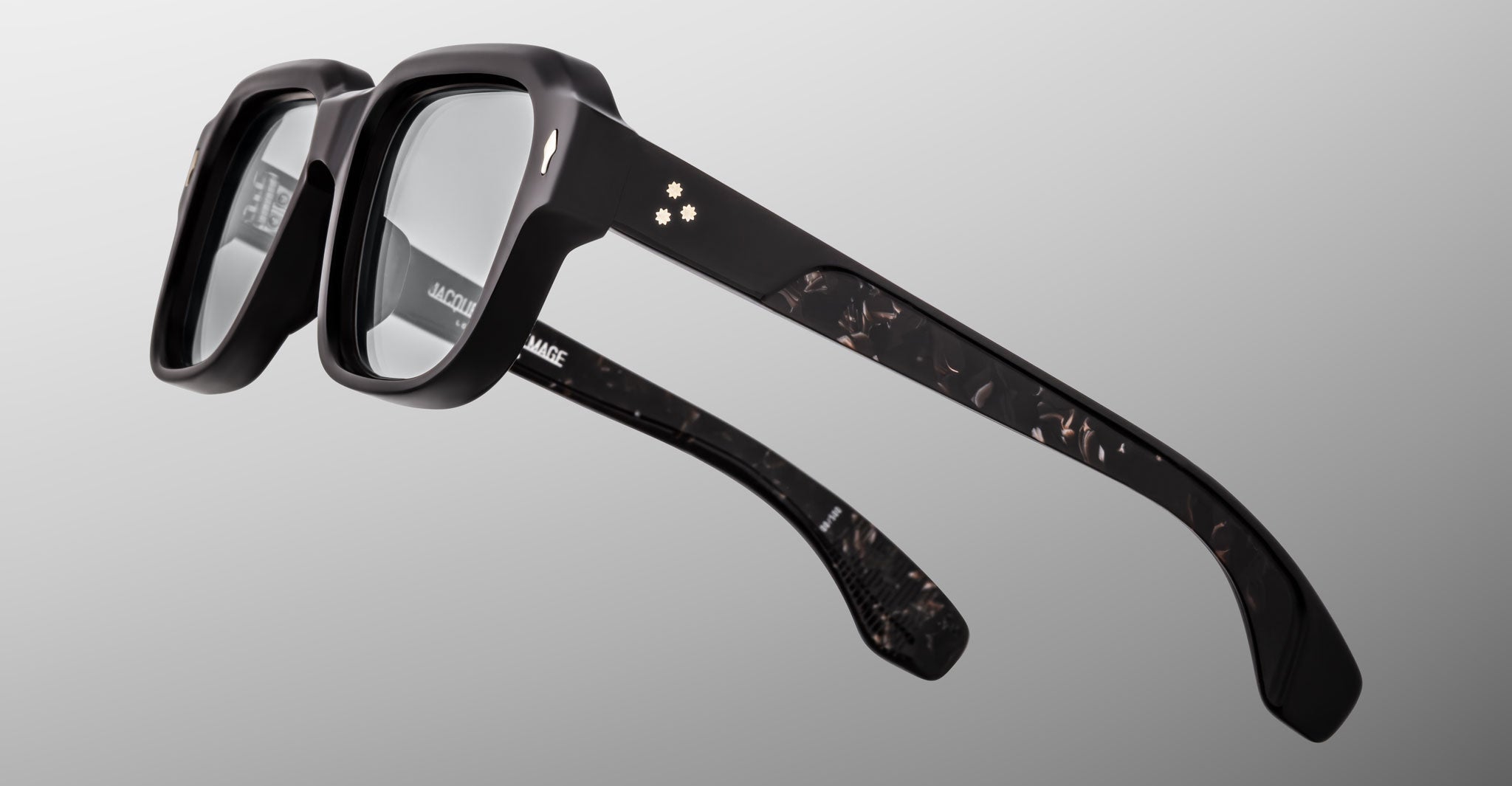 The Jacques Marie Mage Vivienne eyeglasses feature thick, glossy black frames with marbled translucent temples and two subtle star-shaped accents on the sides, set against a smooth light gray gradient background.