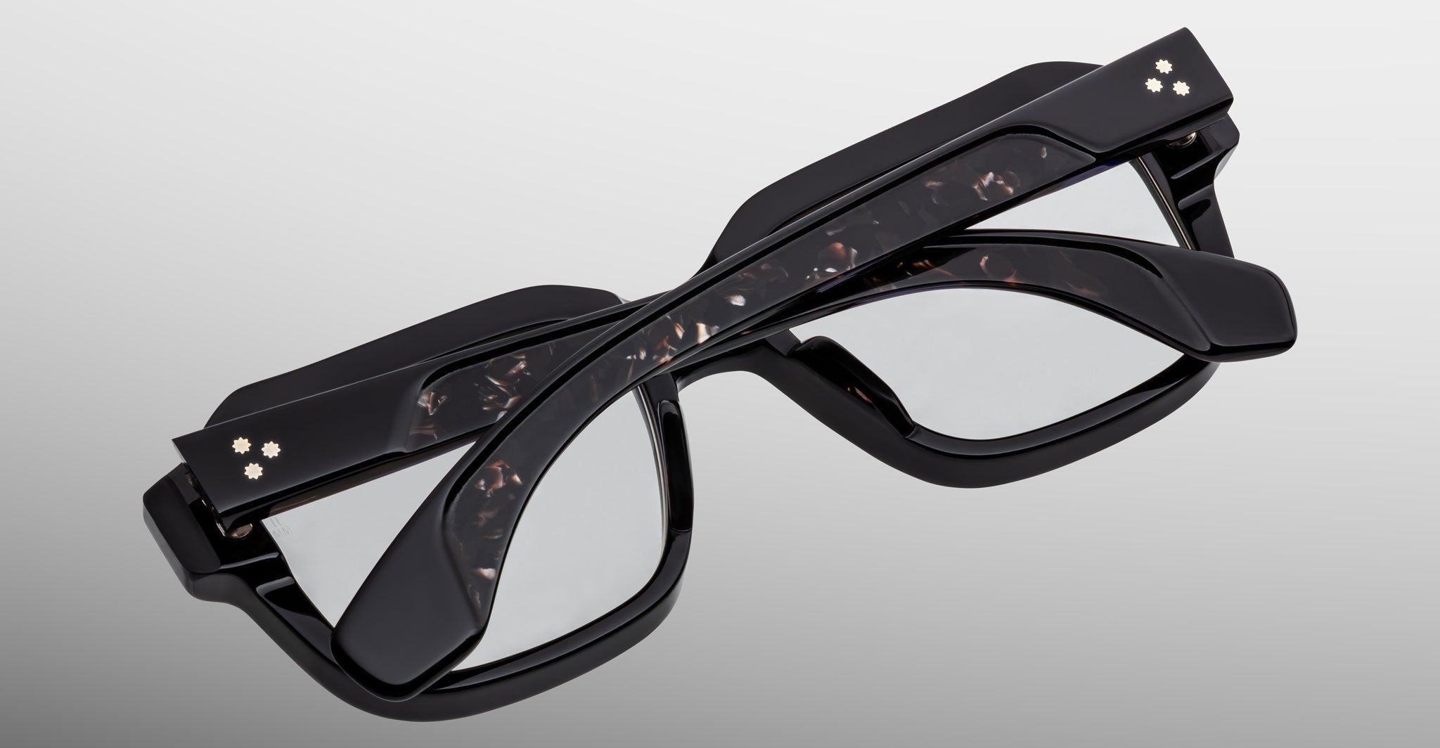 Three Jacques Marie Mage Vivienne eyeglasses with dark, thick frames are stacked together against a plain background. Each pair features two small, flower-like studs on the arms near the hinges.