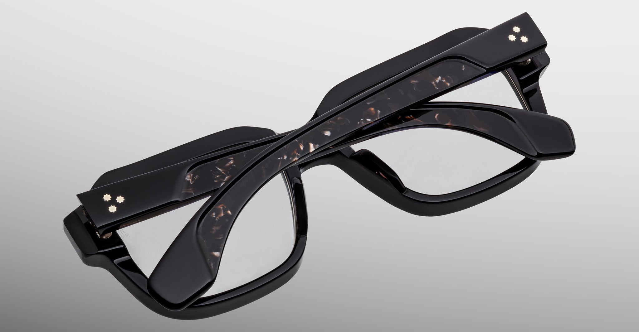 Three Jacques Marie Mage Vivienne eyeglasses with dark, thick frames are stacked together against a plain background. Each pair features two small, flower-like studs on the arms near the hinges.