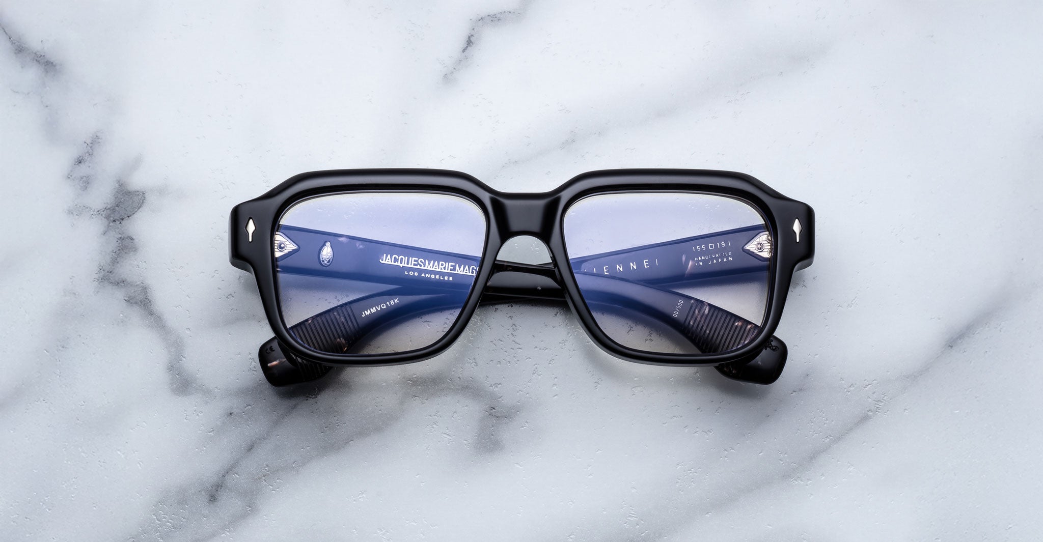 Jacques Marie Mage Vivienne black rectangular eyeglasses with thick frames and blue-tinted lenses, displayed on a white marble surface with the arms folded inward.