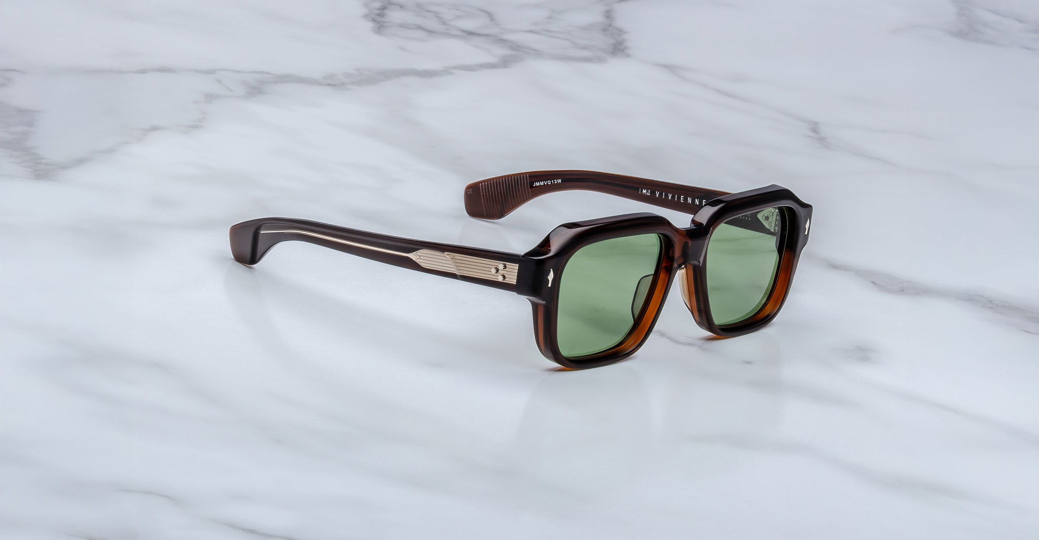 Jacques Marie Mage Vivienne rectangular sunglasses with green-tinted lenses, thick dark brown frames, and metallic temple accents rest on a white marble surface.