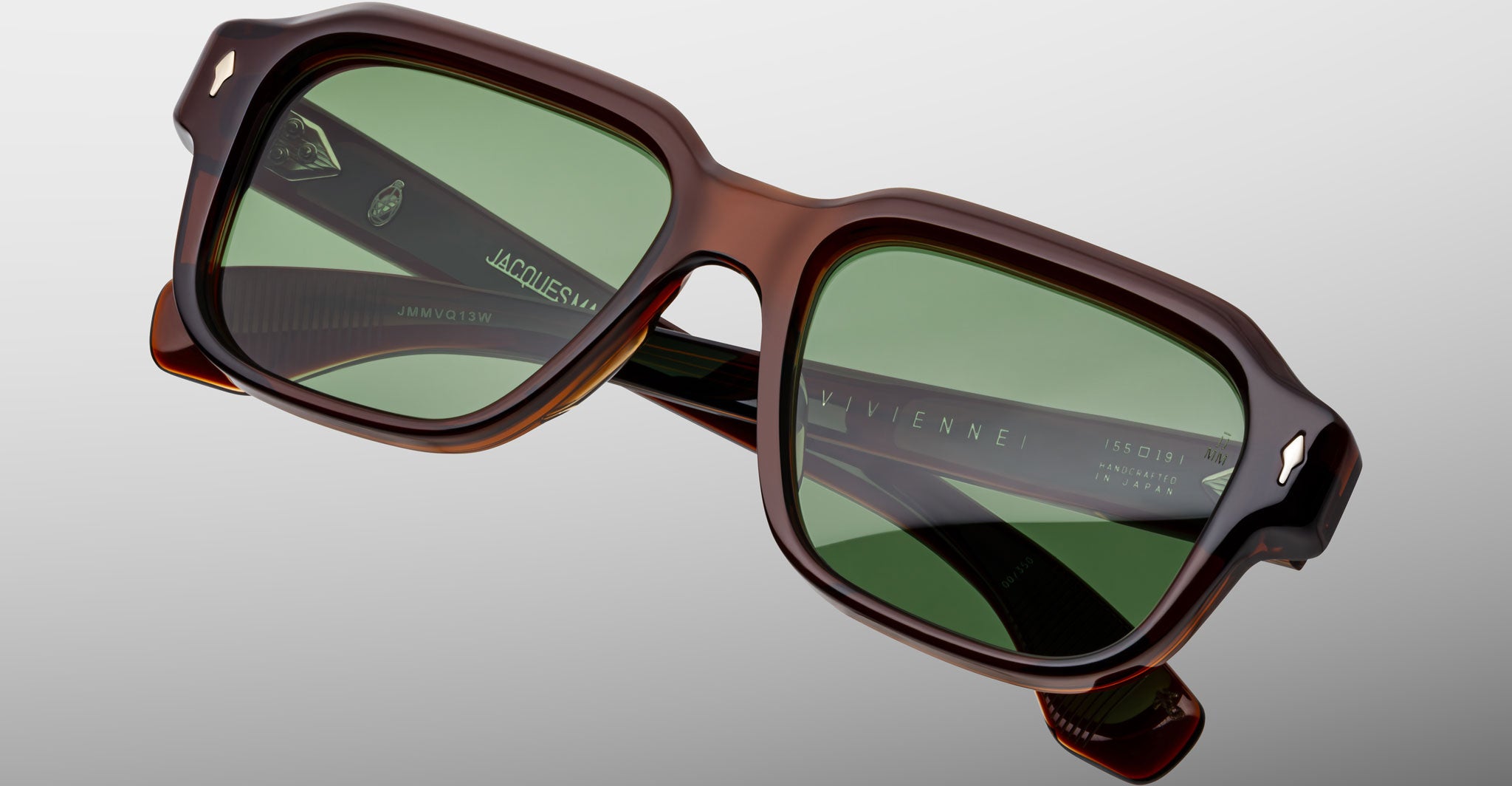 Close-up of Jacques Marie Mage Vivienne brown rectangular sunglasses featuring green-tinted lenses, detailed frame design, and reflective glass, set against a light gray background.