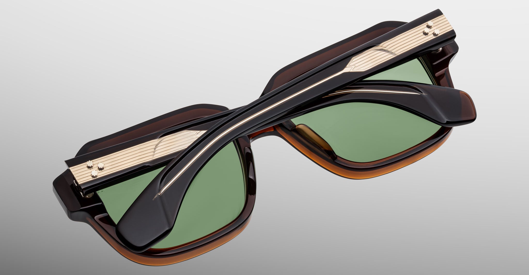 The Jacques Marie Mage Vivienne sunglasses feature green lenses, black and brown frames, and gold accents, displayed folded with crossed arms on a light gray background.