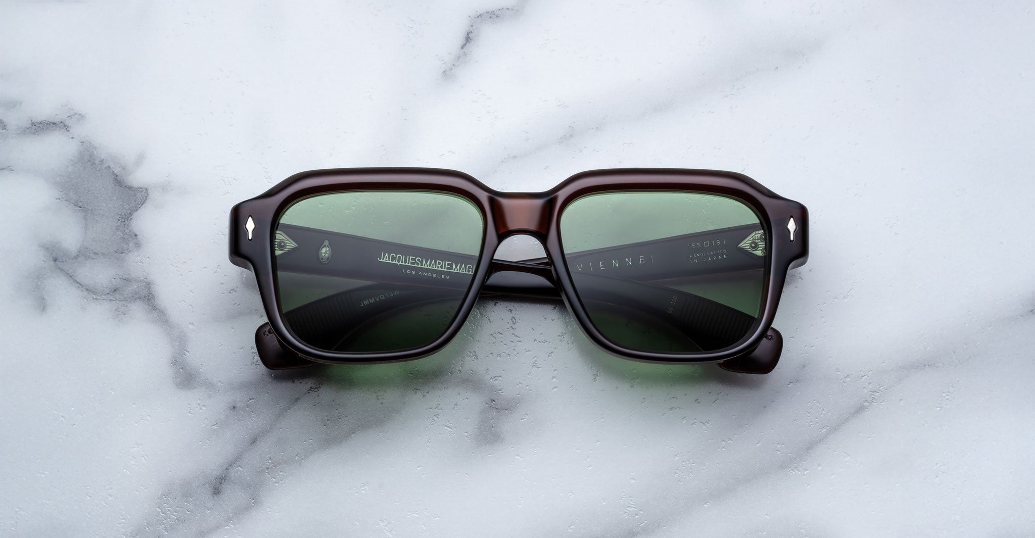 A pair of Jacques Marie Mage Vivienne sunglasses with square dark frames and green-tinted lenses rests on a white marble surface.