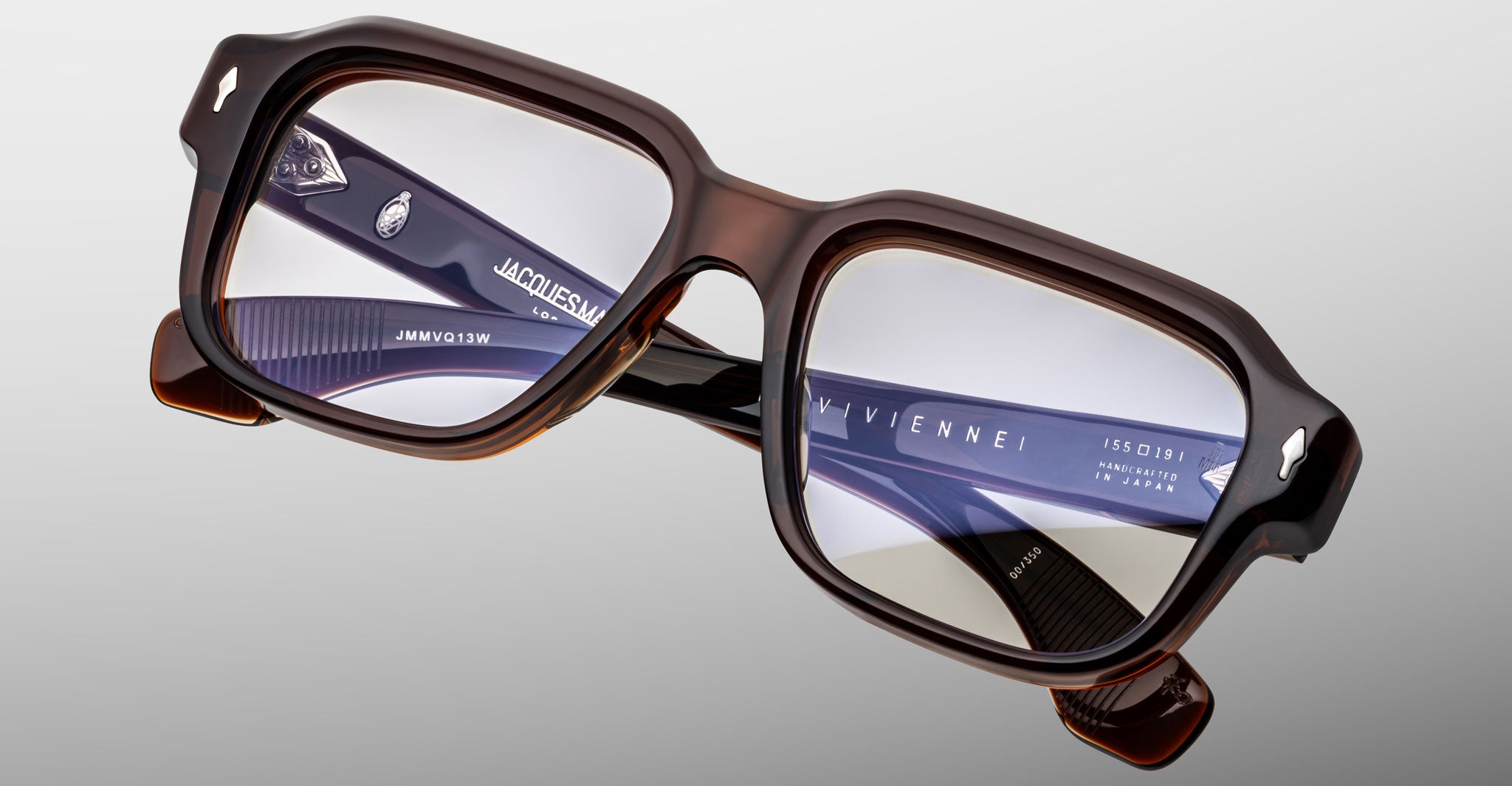 Close-up of Jacques Marie Mage Vivienne eyeglasses: stylish rectangular brown frames with thick edges and dark arms featuring "JACQUES MARIE MAGE" and "VIVIENNE" in white lettering, set against a light gray background.