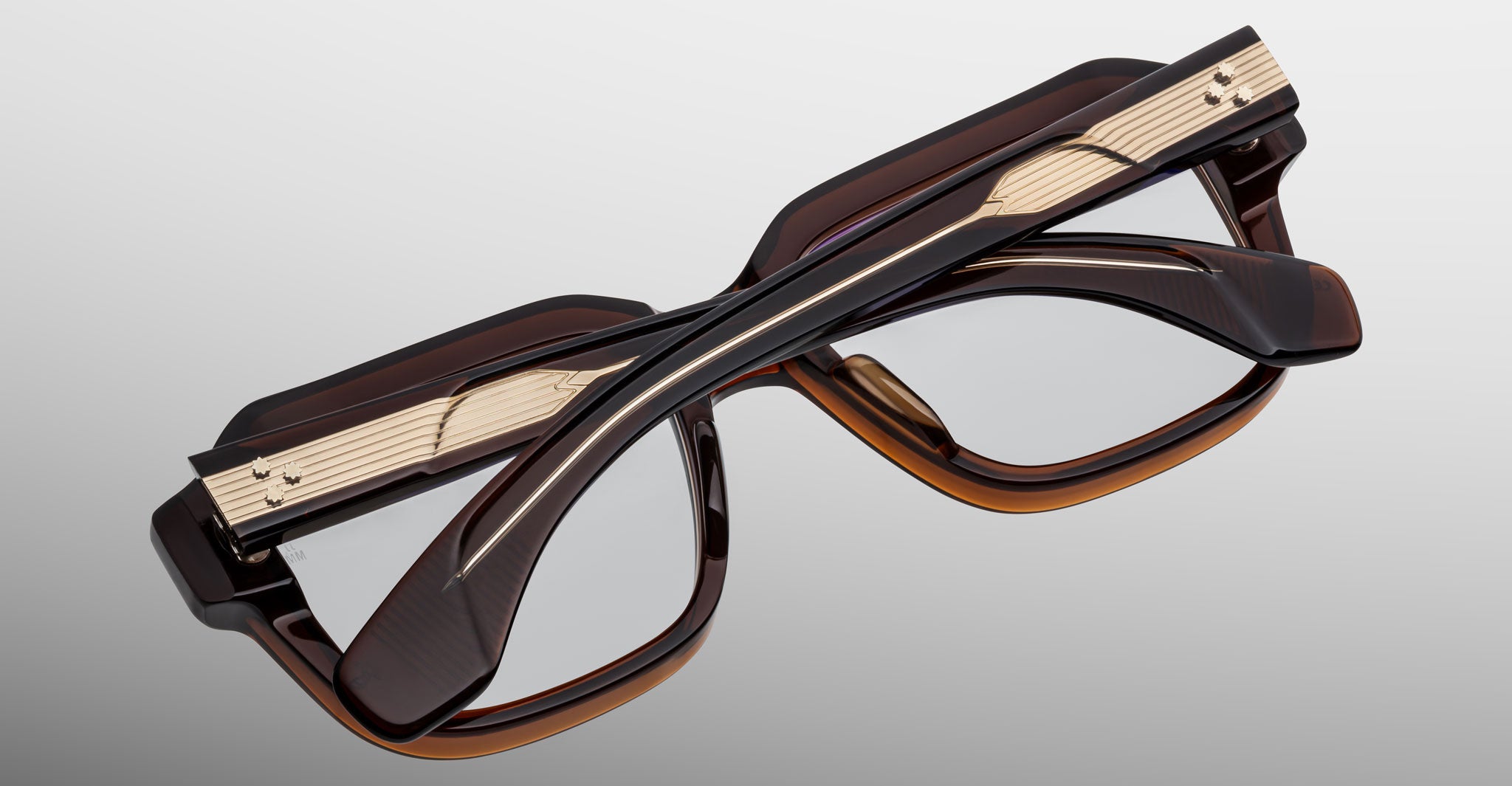 The Jacques Marie Mage Vivienne eyeglasses feature thick dark brown frames with metallic gold accents, layered details, and a modern geometric shape, neatly folded on a light gray surface.
