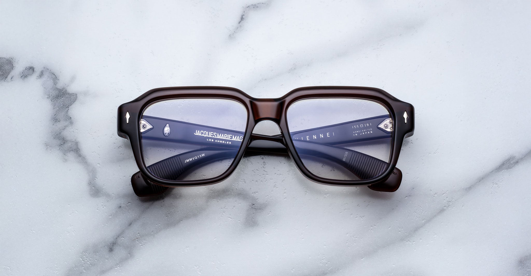 A pair of Jacques Marie Mage Vivienne rectangular sunglasses with dark frames and slightly tinted lenses rests on a white marble surface.