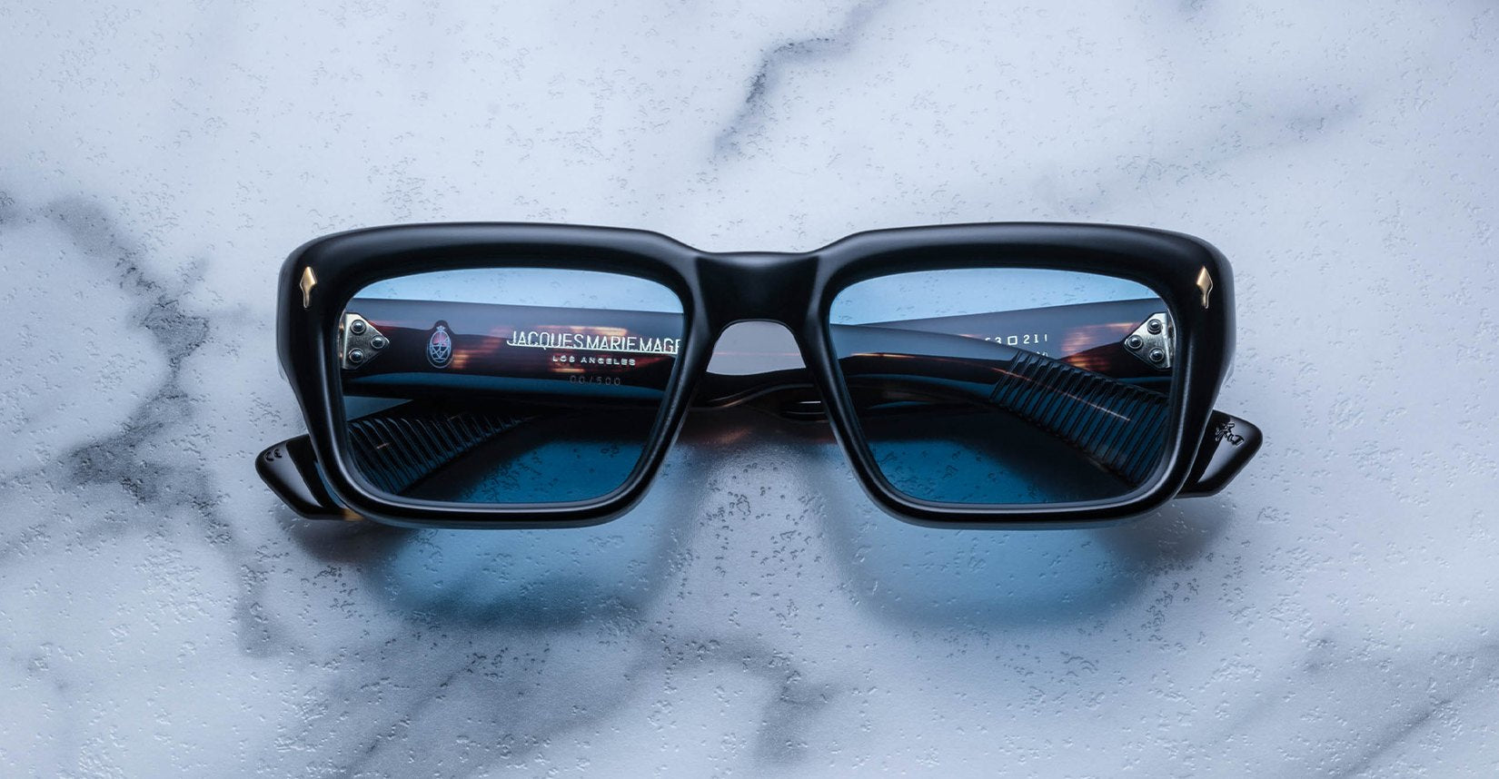 A pair of Jacques Marie Mage Walker black square-framed sunglasses with blue lenses rests on a white marble surface, reflecting a partial logo and inner arm text visible through the lenses.