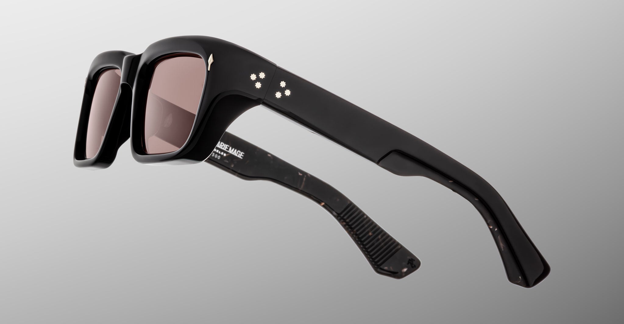 The Jacques Marie Mage Walker sunglasses feature thick rectangular black frames, dark tinted lenses, and small star-shaped accents on the arms, shown against a light gray gradient background.