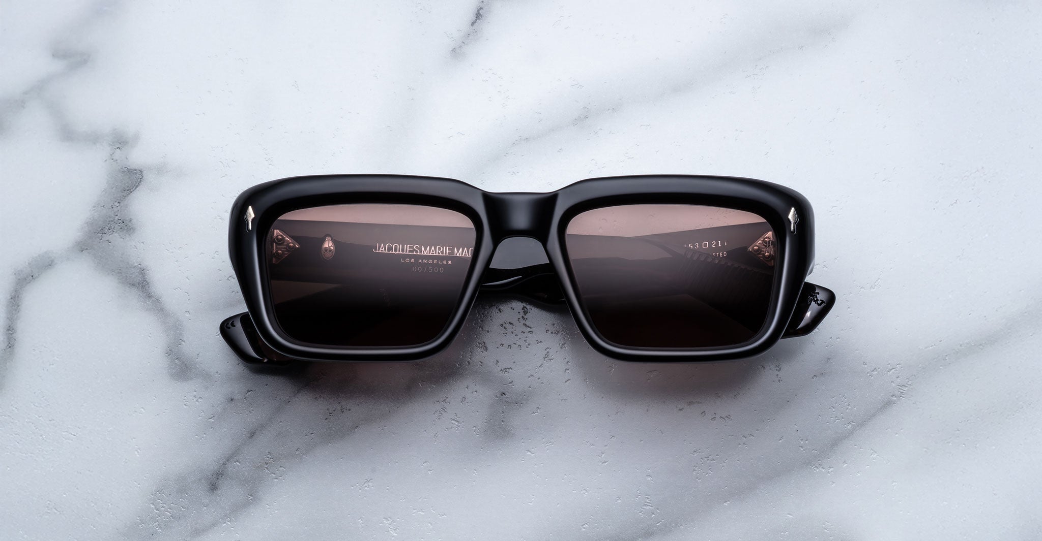 Jacques Marie Mage Walker black square-frame sunglasses with dark lenses rest on a white marble surface, displaying reflections of text and a light source on the lenses.