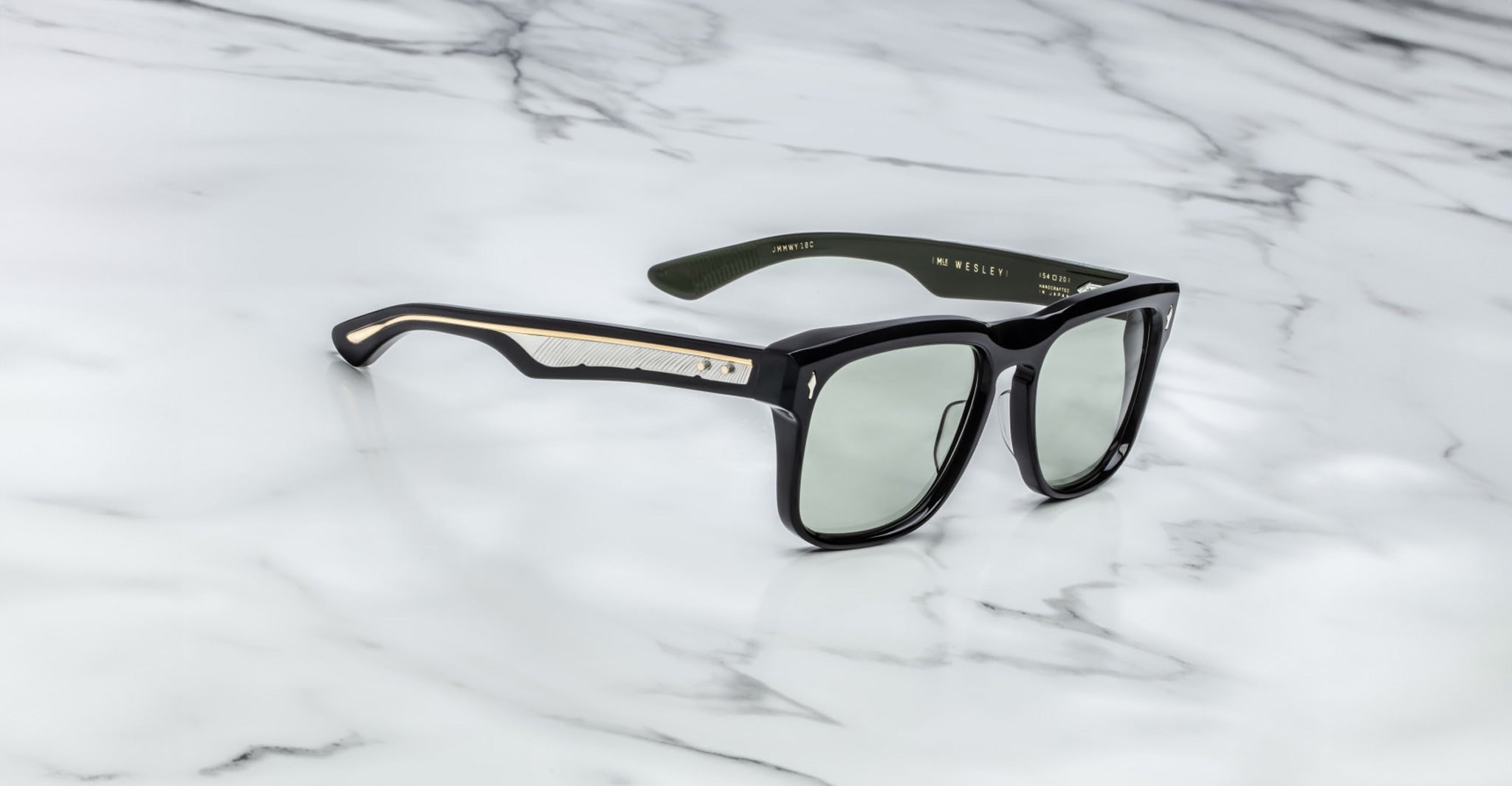 The Jacques Marie Mage Wesley sunglasses, featuring black frames, green-tinted lenses, and gold-accented arms, rest on a white marble surface.