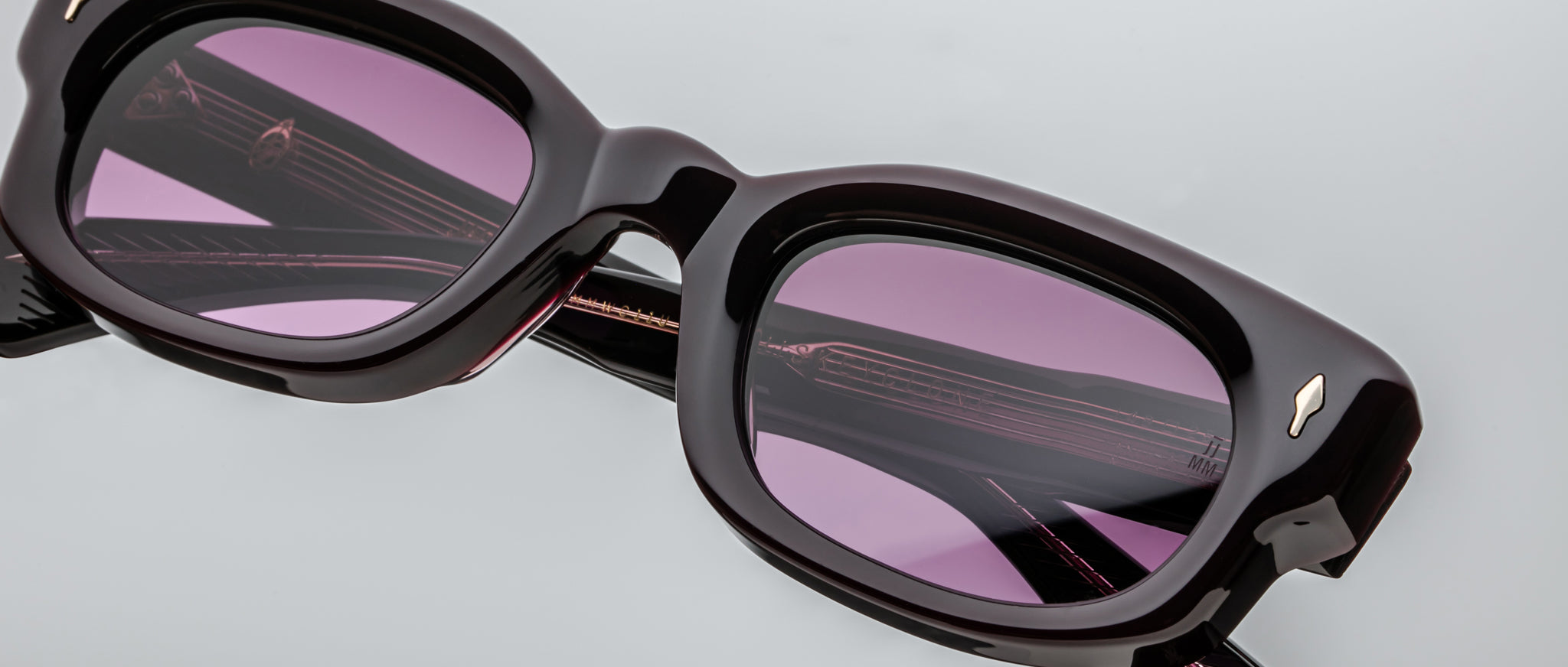 Close-up of Jacques Marie Mage Whiskeyclone black rectangular eyeglasses with purple-tinted lenses, shown against a light gray background. The lenses reflect the glasses’ temples and subtle detailing.