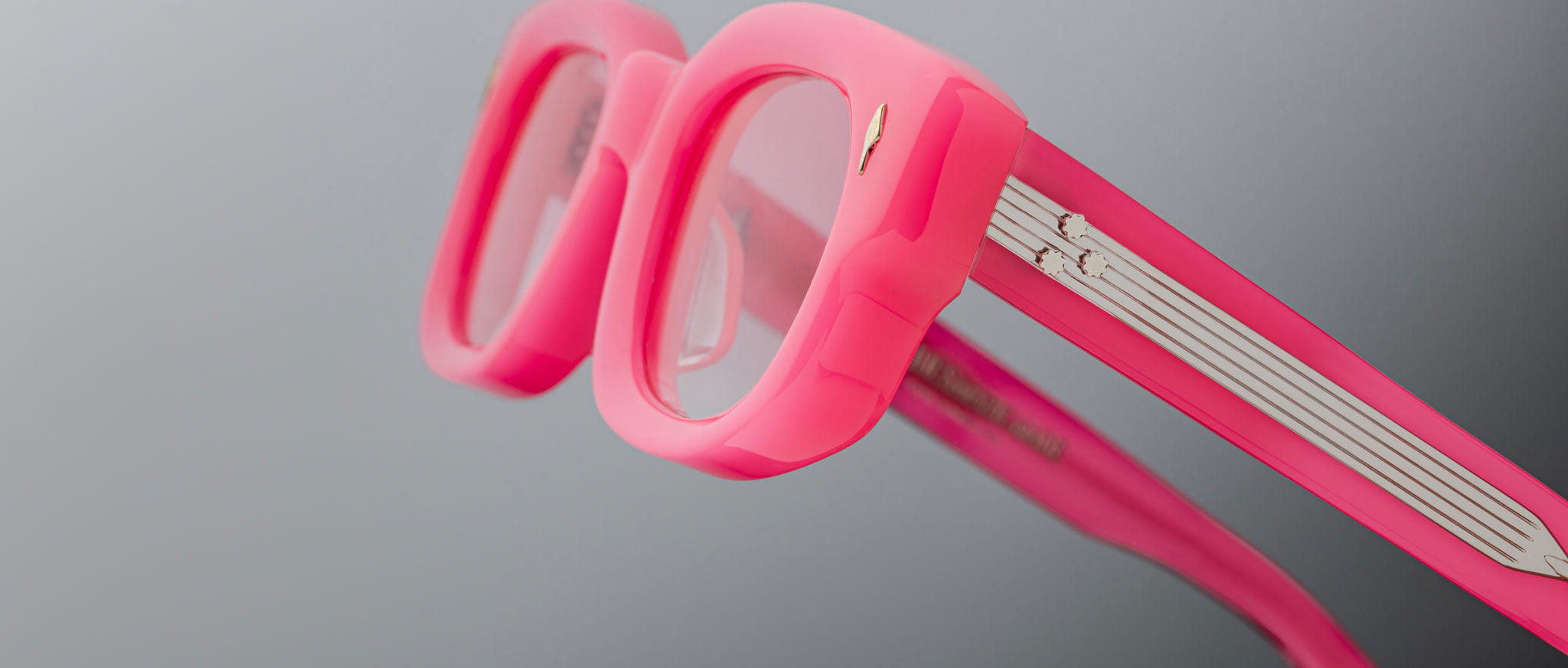 Close-up of Jacques Marie Mage Whiskeyclone eyeglasses featuring thick, bright pink frames and clear lenses against a neutral gray background; temples showcase white and silver detailing.