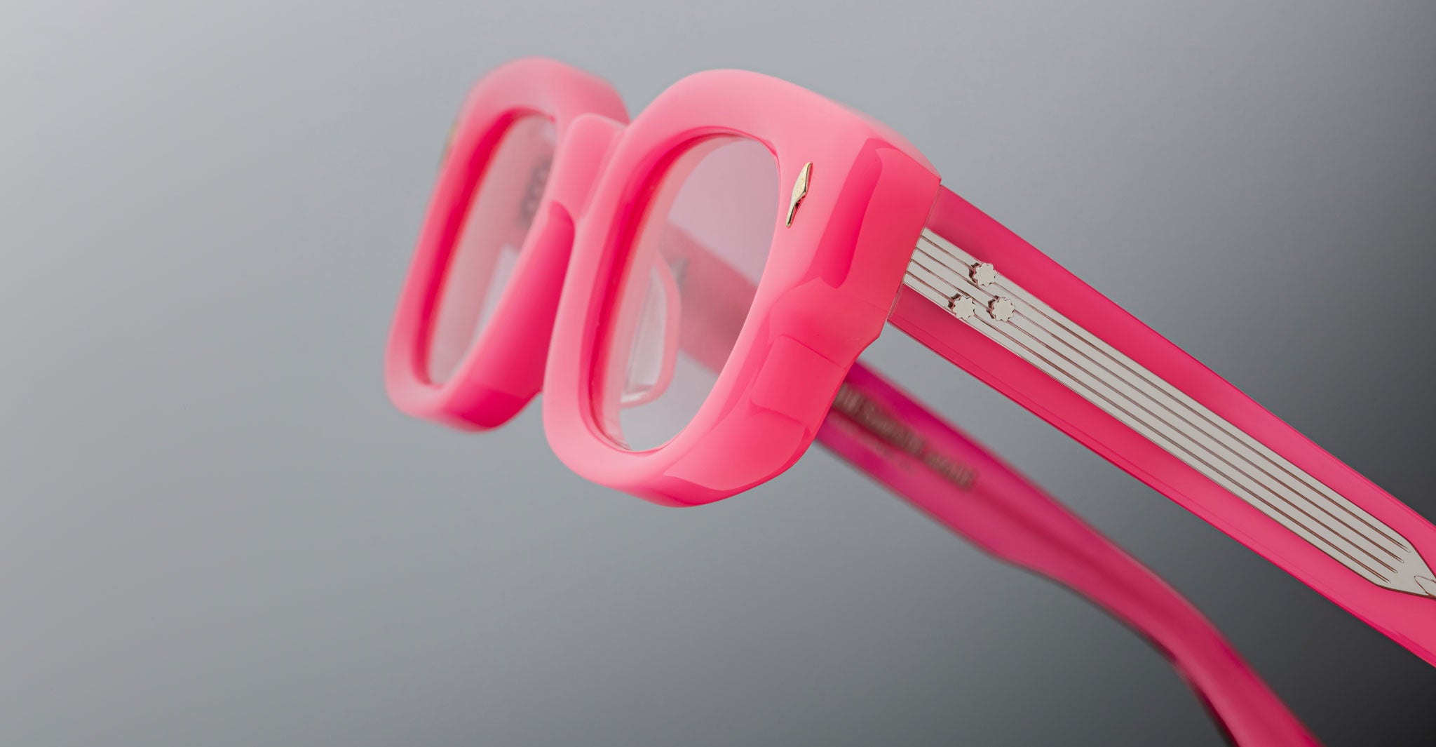 Close-up of Jacques Marie Mage Whiskeyclone eyeglasses featuring thick, bright pink frames and clear lenses against a neutral gray background; temples showcase white and silver detailing.