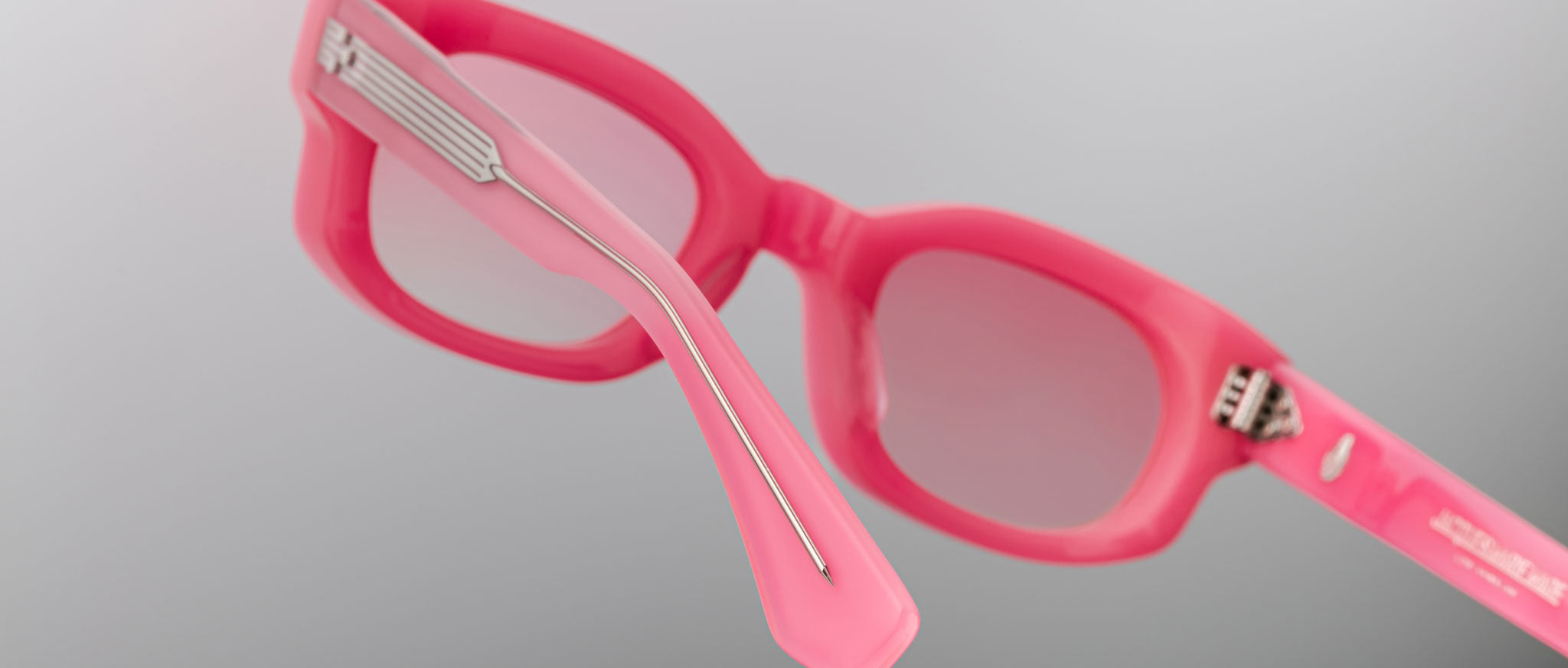 Close-up of Jacques Marie Mage Whiskeyclone sunglasses with thick pink frames and pink-tinted lenses. The plain light gray background highlights the prominent left temple arm with a silver metal detail.