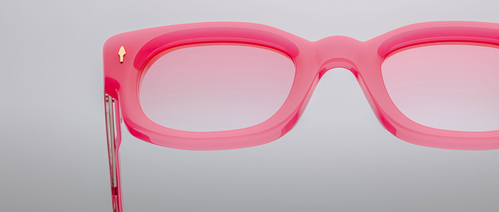 Close-up of the right side of Jacques Marie Mage Whiskeyclone eyeglass frames in pink with matching lenses. A small gold arrow detail appears near the hinge. Set against a plain, light gray background.