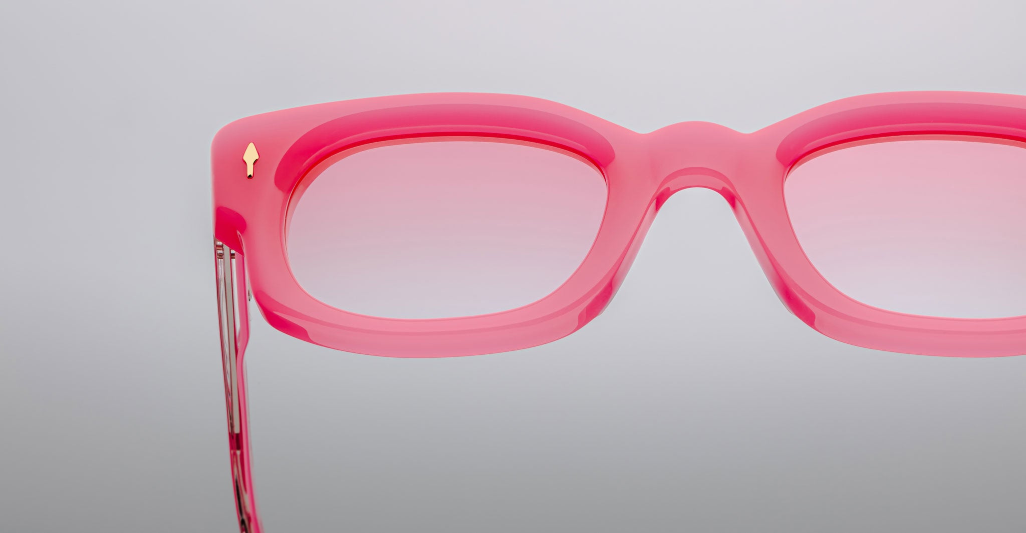 Close-up of the right side of Jacques Marie Mage Whiskeyclone eyeglass frames in pink with matching lenses. A small gold arrow detail appears near the hinge. Set against a plain, light gray background.
