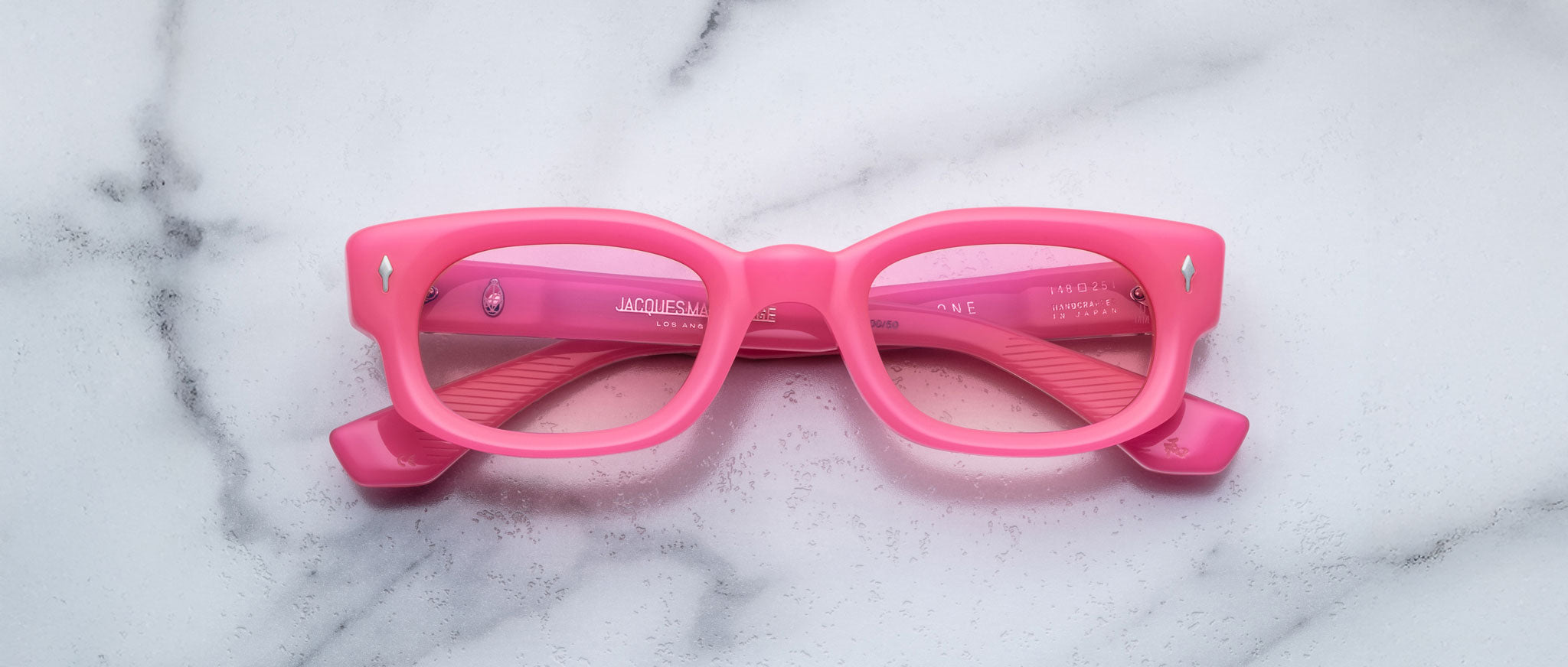 The Jacques Marie Mage Whiskeyclone features bright pink rectangular frames with slightly thick rims, displayed facing up on a white marble surface with subtle gray veins and inward-folded temples.