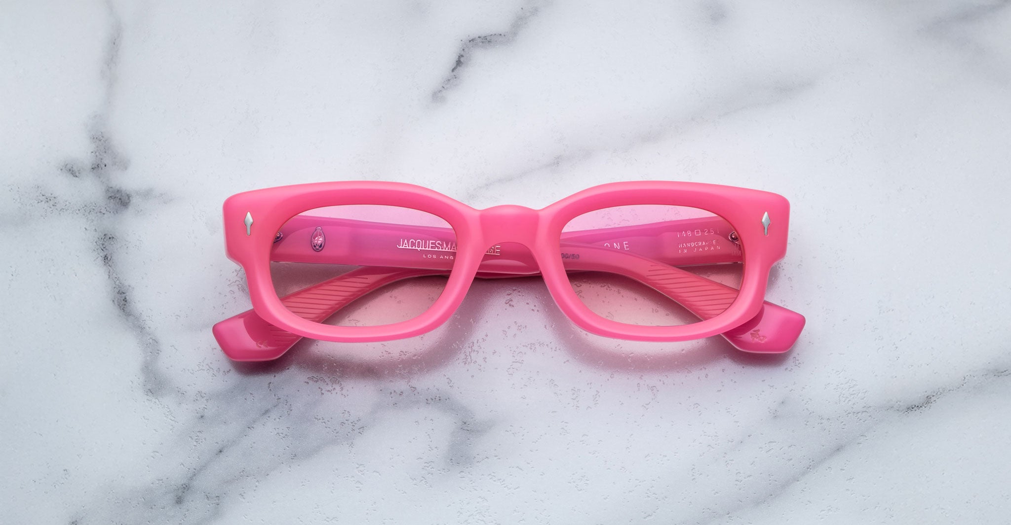 The Jacques Marie Mage Whiskeyclone features bright pink rectangular frames with slightly thick rims, displayed facing up on a white marble surface with subtle gray veins and inward-folded temples.