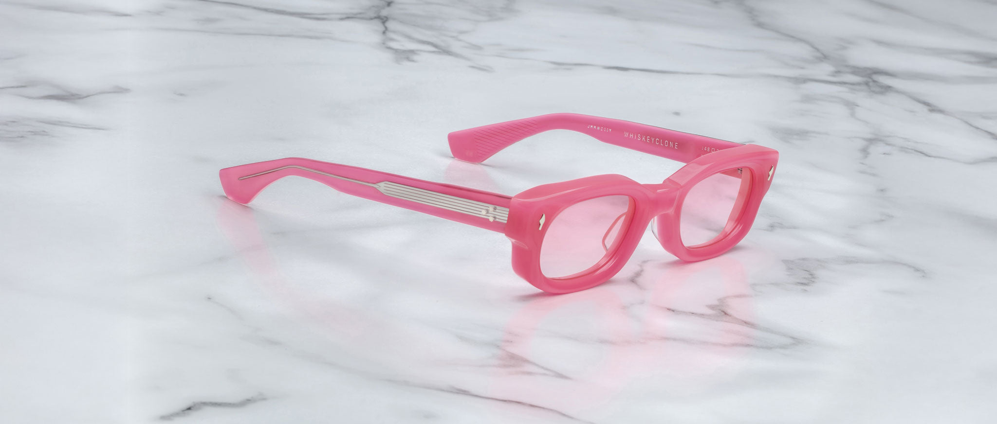 Jacques Marie Mage Whiskeyclone eyeglasses with pink thick rims and arms rest on a white marble surface with gray veins. The arms feature silver horizontal detailing near the hinges.