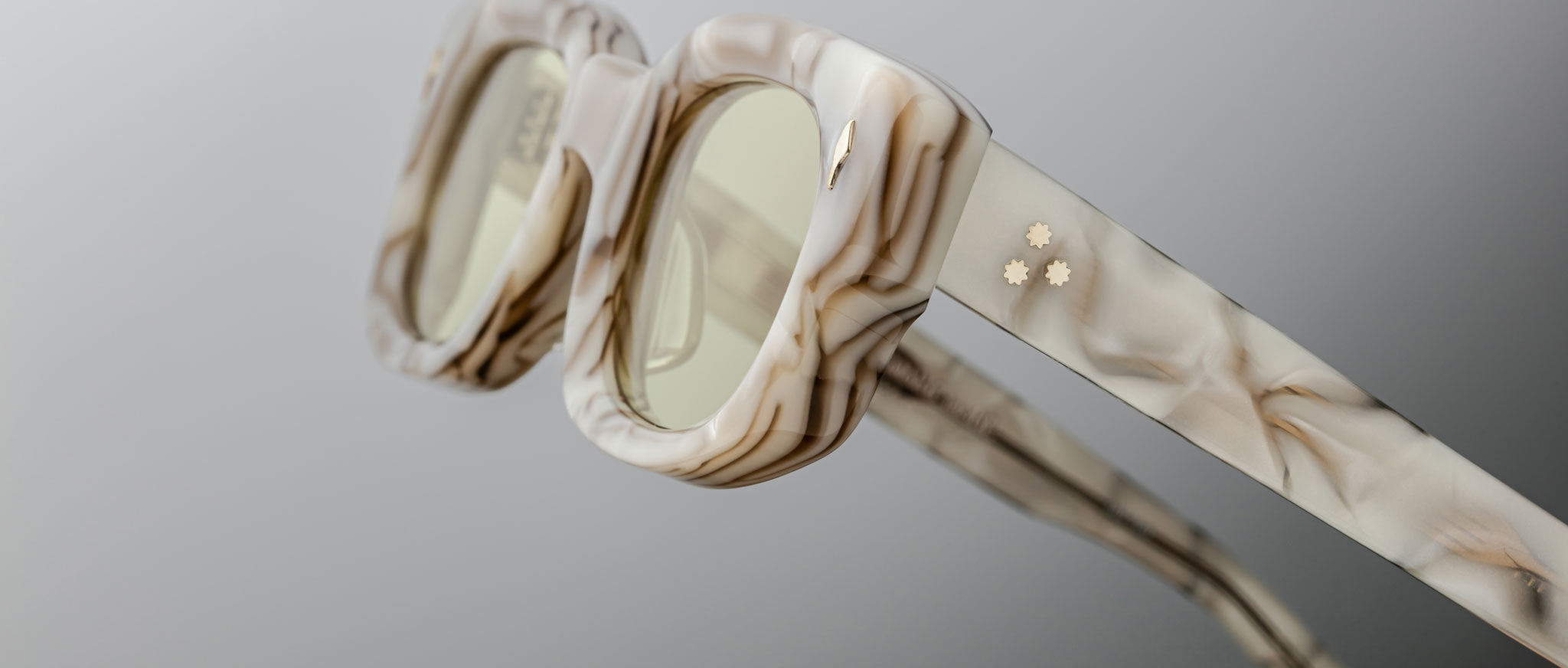 A close-up of Jacques Marie Mage Whiskeyclone square-framed sunglasses featuring a marble pattern, light-tinted lenses, and small gold star-shaped temple details set against a neutral background.