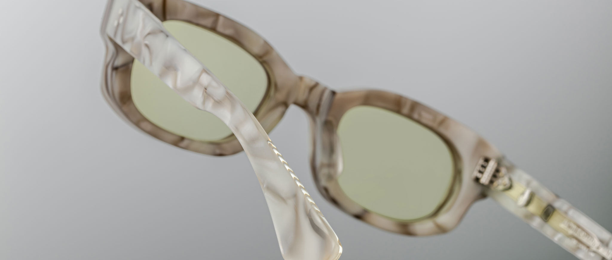 Close-up view of Jacques Marie Mage Whiskeyclone eyeglasses with translucent, marbled frames and light green-tinted lenses, highlighting the frame’s detailed texture against a plain gray background.