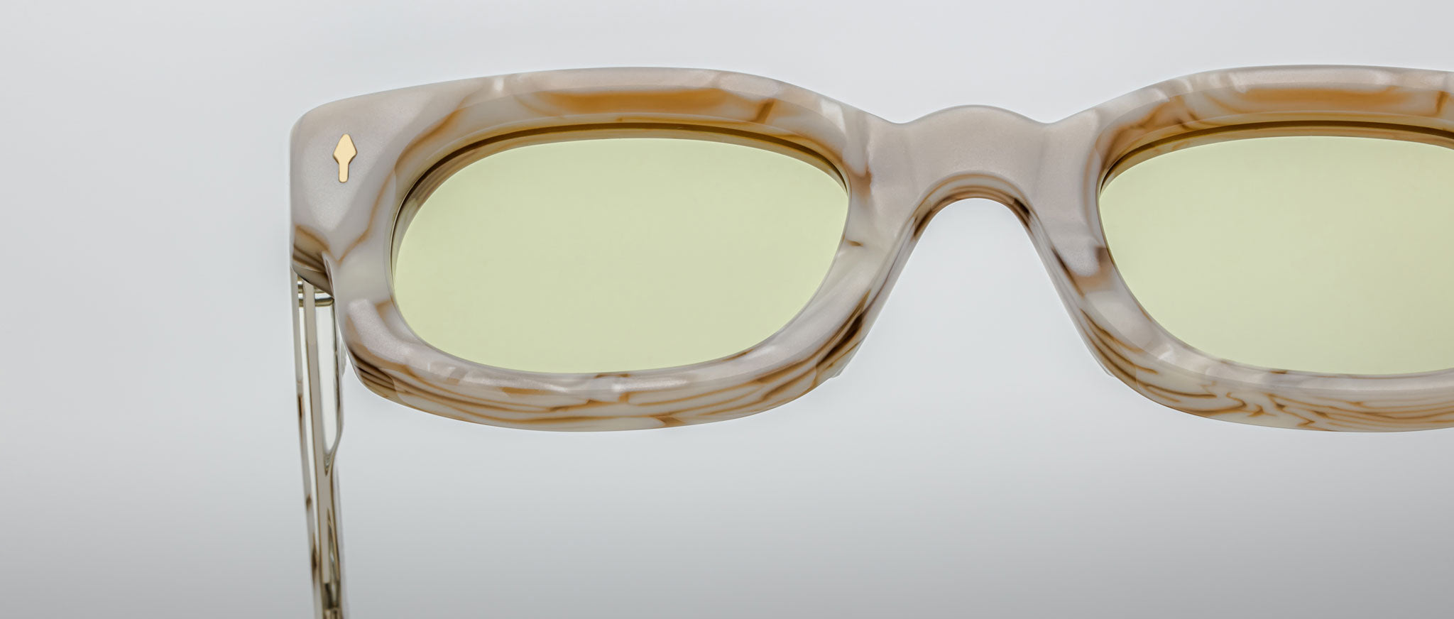 Close-up of Jacques Marie Mage Whiskeyclone rectangular eyeglasses with a marble-patterned frame, light yellow-tinted lenses, and a gold accent, set against a plain light gray background.
