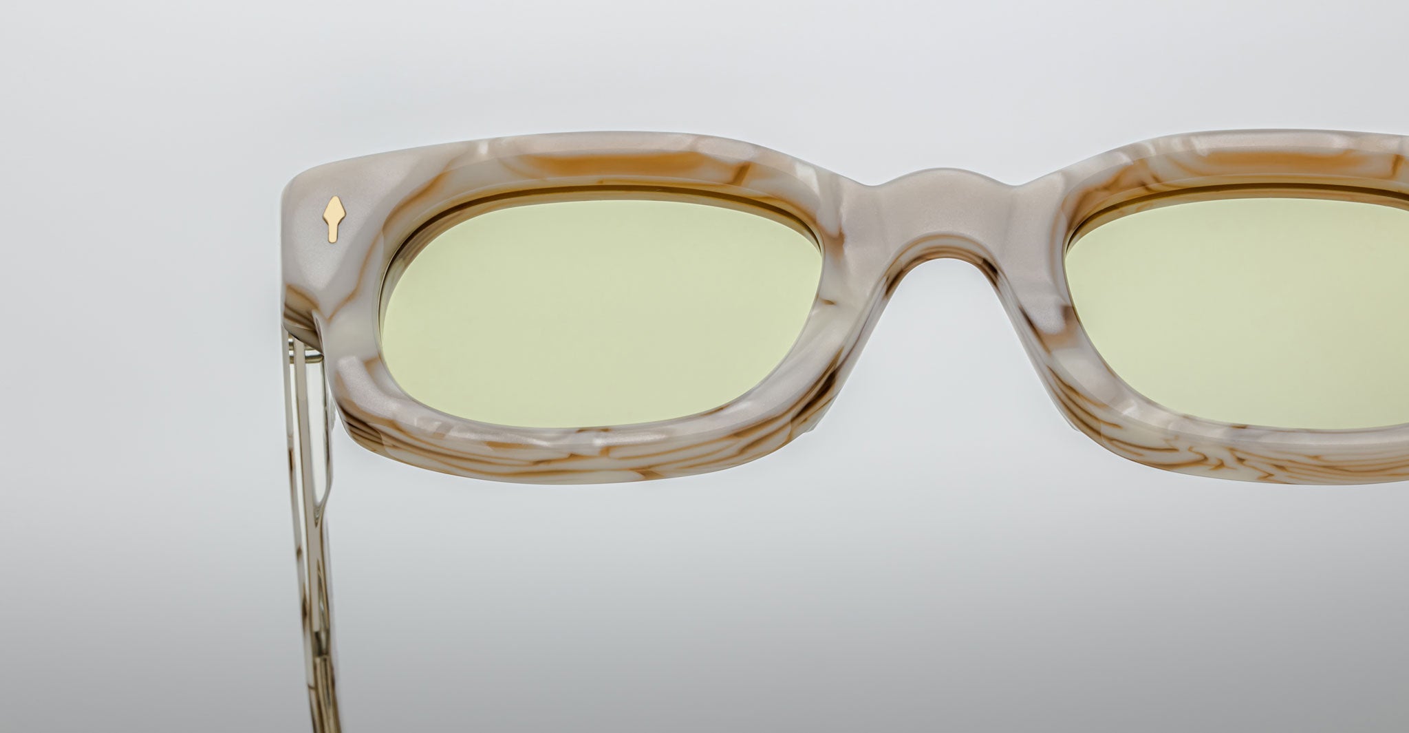 Close-up of Jacques Marie Mage Whiskeyclone rectangular eyeglasses with a marble-patterned frame, light yellow-tinted lenses, and a gold accent, set against a plain light gray background.
