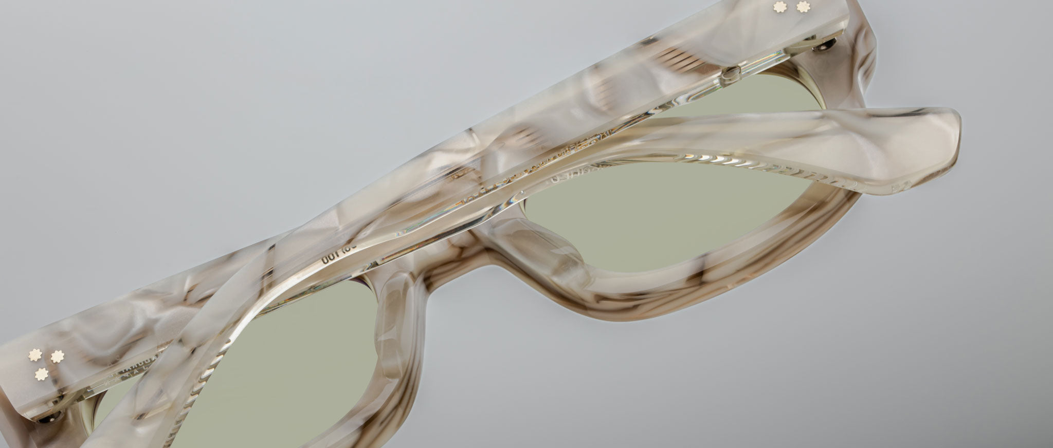 A close-up of Jacques Marie Mage Whiskeyclone rectangular sunglasses with a marbled beige and brown frame, light green lenses, and two small gold flower-shaped studs on one arm, set against a plain gray background.