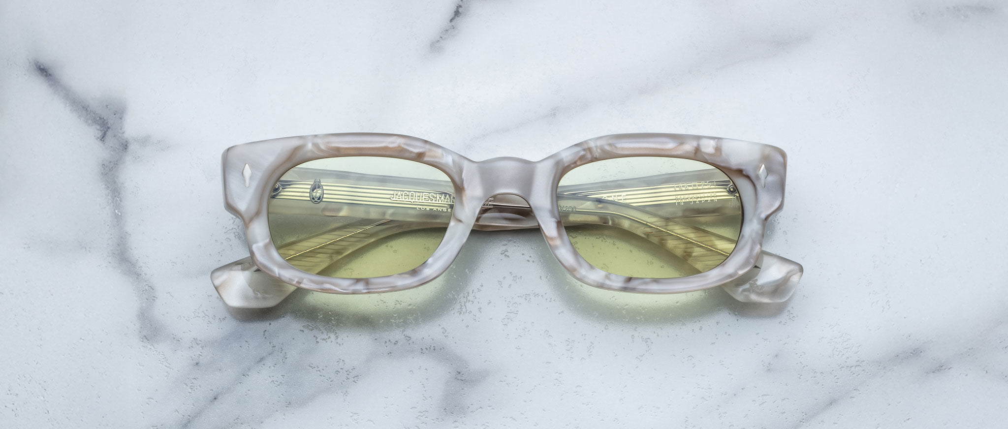 A pair of Jacques Marie Mage Whiskeyclone rectangular eyeglasses with a marble pattern and yellow-tinted lenses rests on a white marble surface.