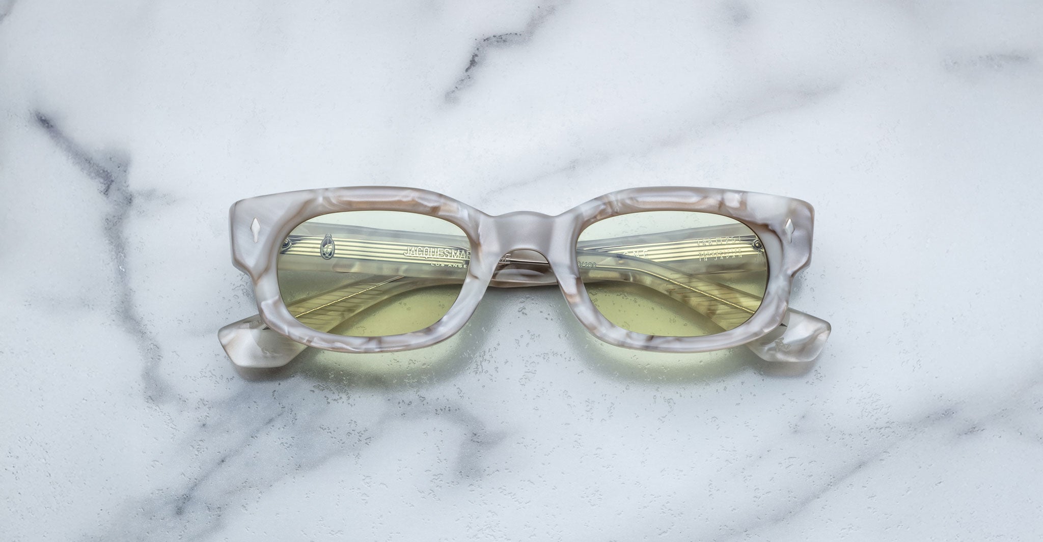 A pair of Jacques Marie Mage Whiskeyclone rectangular eyeglasses with a marble pattern and yellow-tinted lenses rests on a white marble surface.