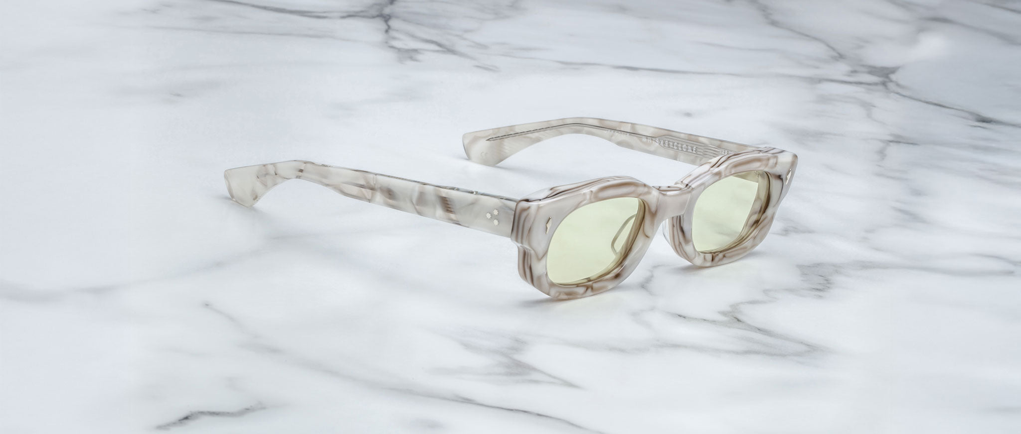 The Jacques Marie Mage Whiskeyclone sunglasses feature a clear marble-patterned frame and arms with a translucent, milky look, paired with light yellow tinted lenses, and are displayed on a white marble surface.